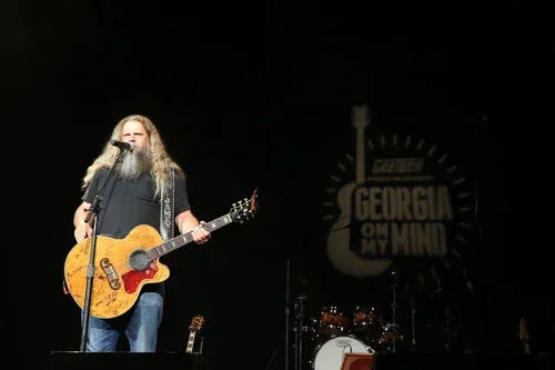 A man with long hair and a beard playing an acoustic guitar on stage, with a microphone in front of him and a drum set behind him.