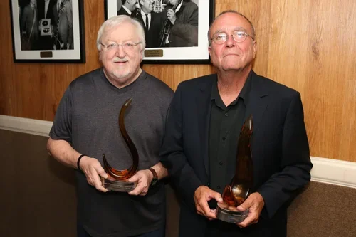 Two men holding award trophies in front of a wooden wall with black and white photographs, smiling at the camera.