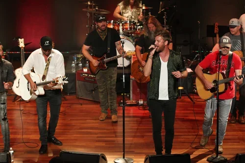 A band performing on stage with five members playing guitars, one member singing into a microphone, and a drummer in the background.