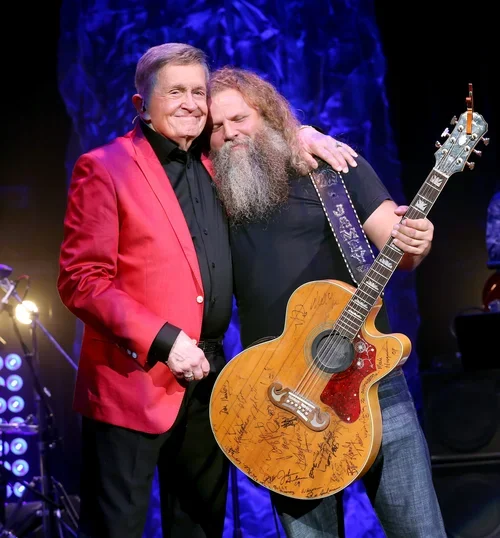 Two men on stage, one wearing a red blazer and the other with a beard holding an autographed guitar, hugging each other.