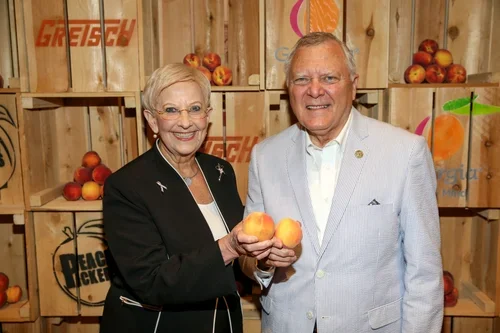 An older woman and man smiling and holding peaches at a display of peaches in wooden crates, behind them are colorful fruit-themed decorations.