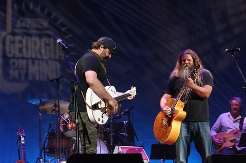 Two musicians playing acoustic guitars on stage, with a band setup including drums and amplifiers, during a live performance.