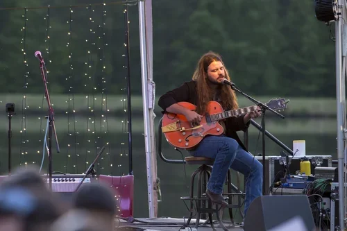Male musician with long hair and beard playing an acoustic guitar on an outdoor stage.