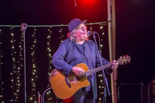 A musician singing into a microphone while playing an acoustic guitar on stage with string lights in the background.