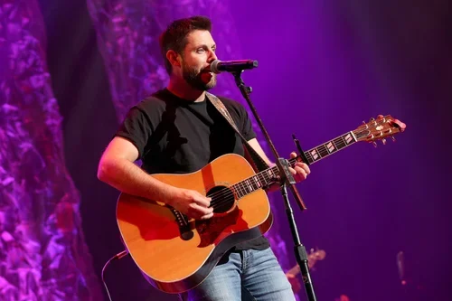 Man with a beard playing an acoustic guitar on stage with purple lighting.