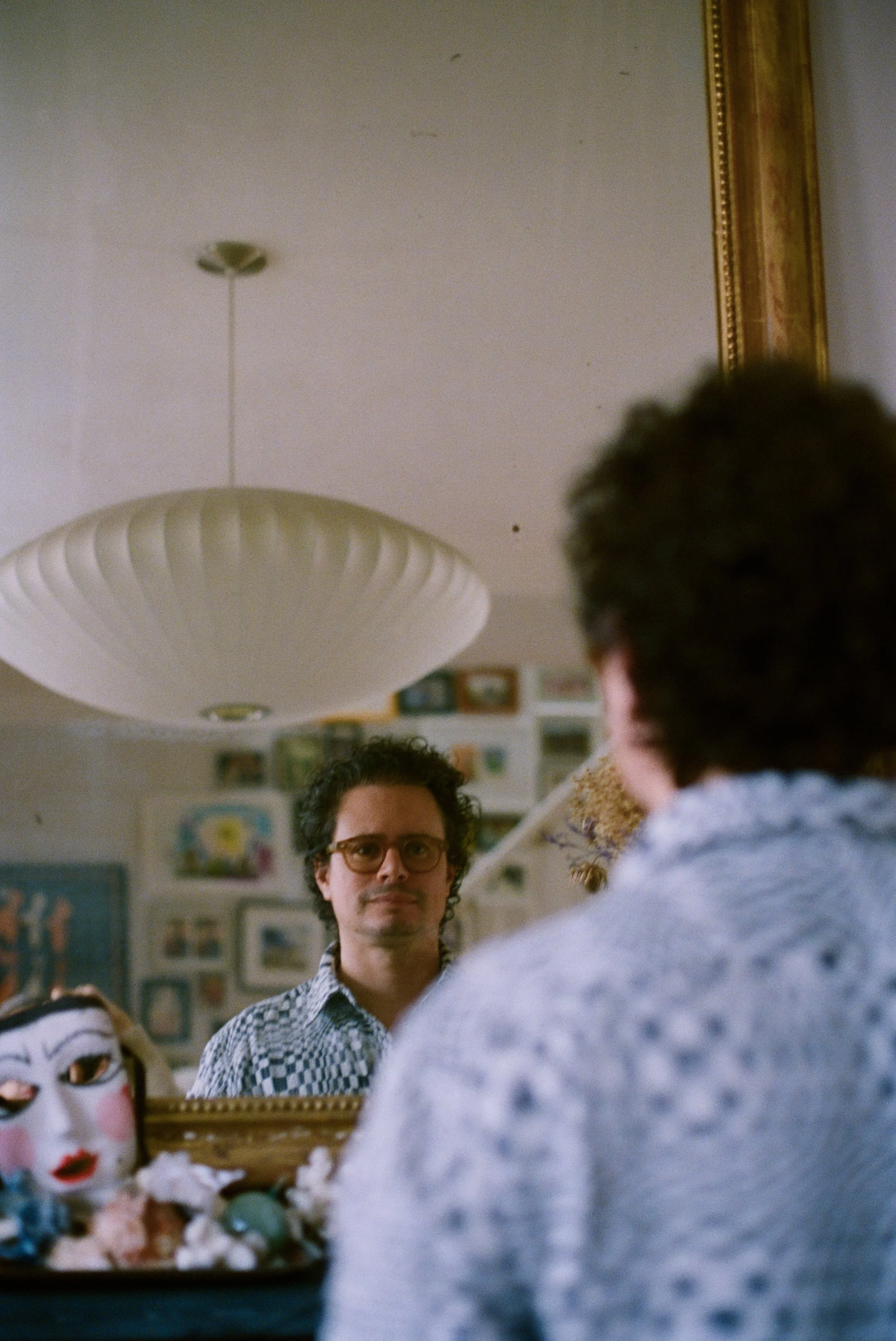 Photo of Nate Martinez. A person with curly hair and glasses looking at a person in front of them who is wearing a patterned shirt, in a room with various framed pictures on the wall, and a mirror with a decorative frame in the background.