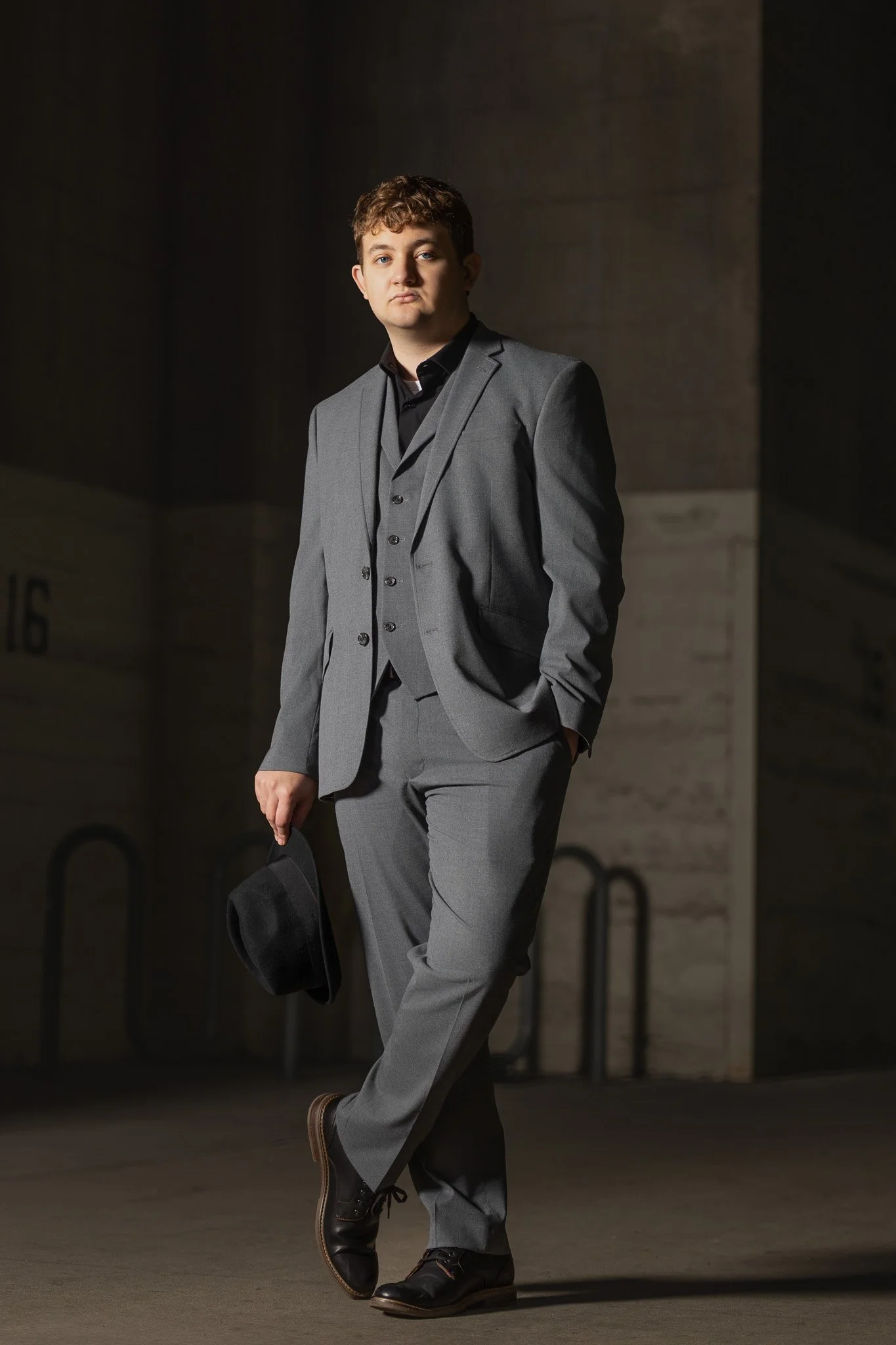 Senior boy at Kansas City Union Station in gray suit holding a black hat, standing outdoors in an urban setting, with a dark background.