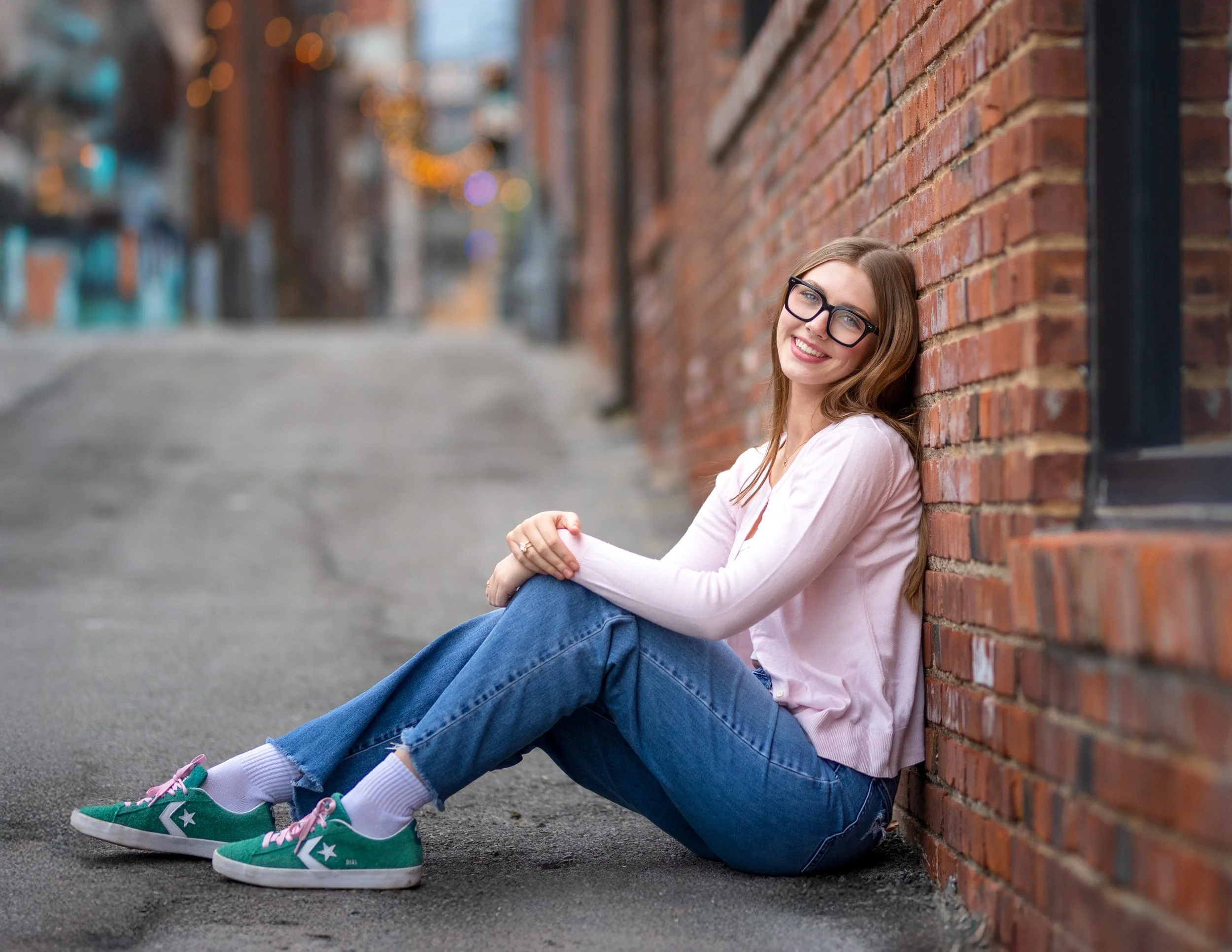 Senior girl leaning against a downtown Kansas City brick wall with long brown hair, glasses, and a bright smile, wearing a pink jacket, jeans, and green sneakers, sitting on the sidewalk 