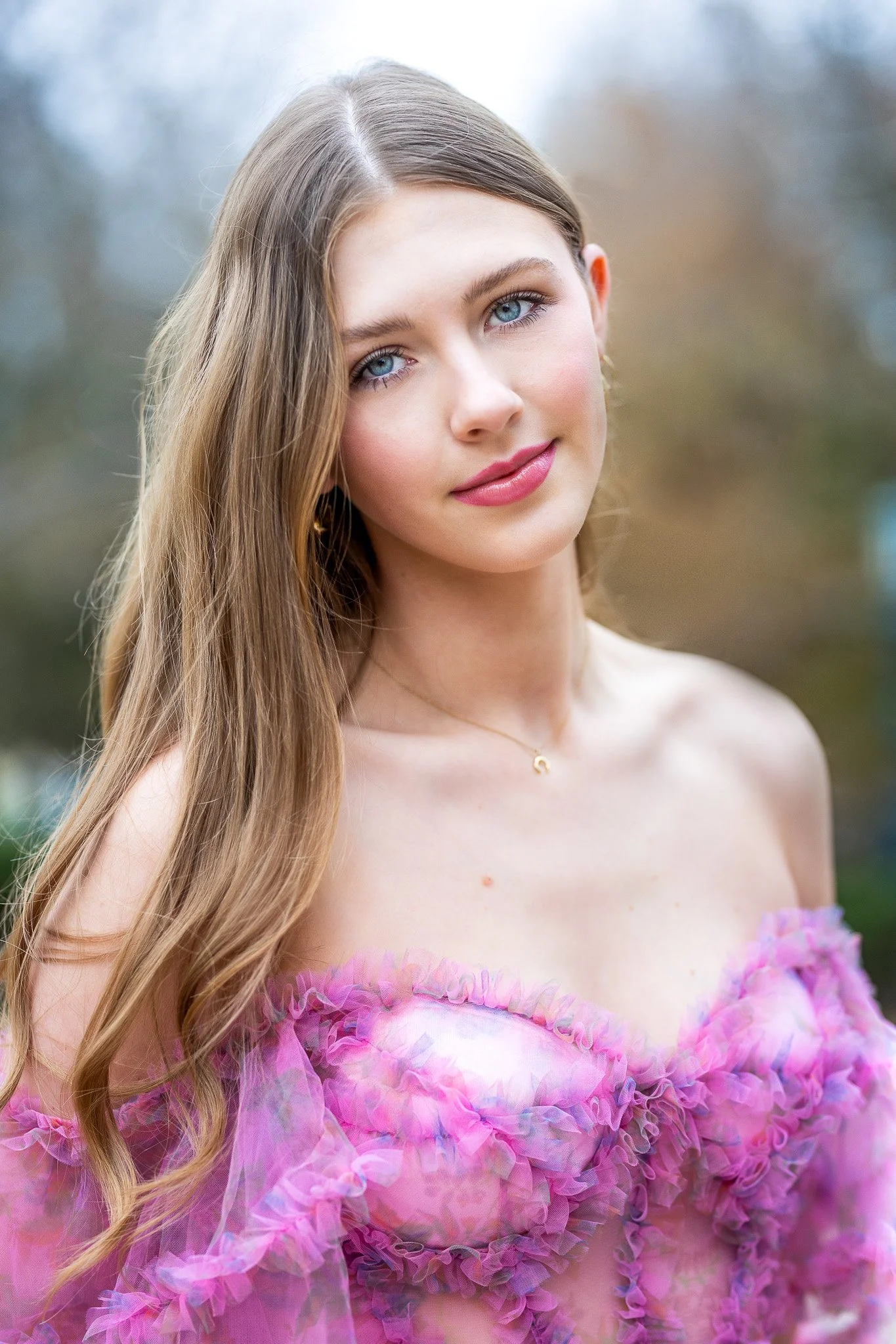 A senior girl with long brown hair, smiling, wearing a red sleeveless dress with thin straps, outdoors with autumn trees in the background.