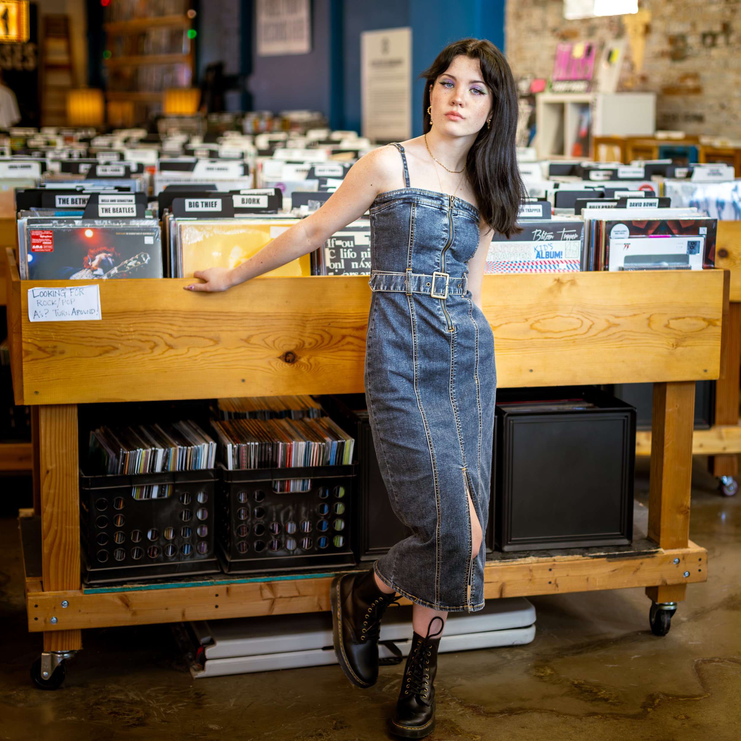Editorial senior photo of a girl wearing a sleeveless denim jumpsuit, standing in a Kansas City record store,  with dark hair and makeup, posing with her hand on a wooden display table filled with vinyl records.