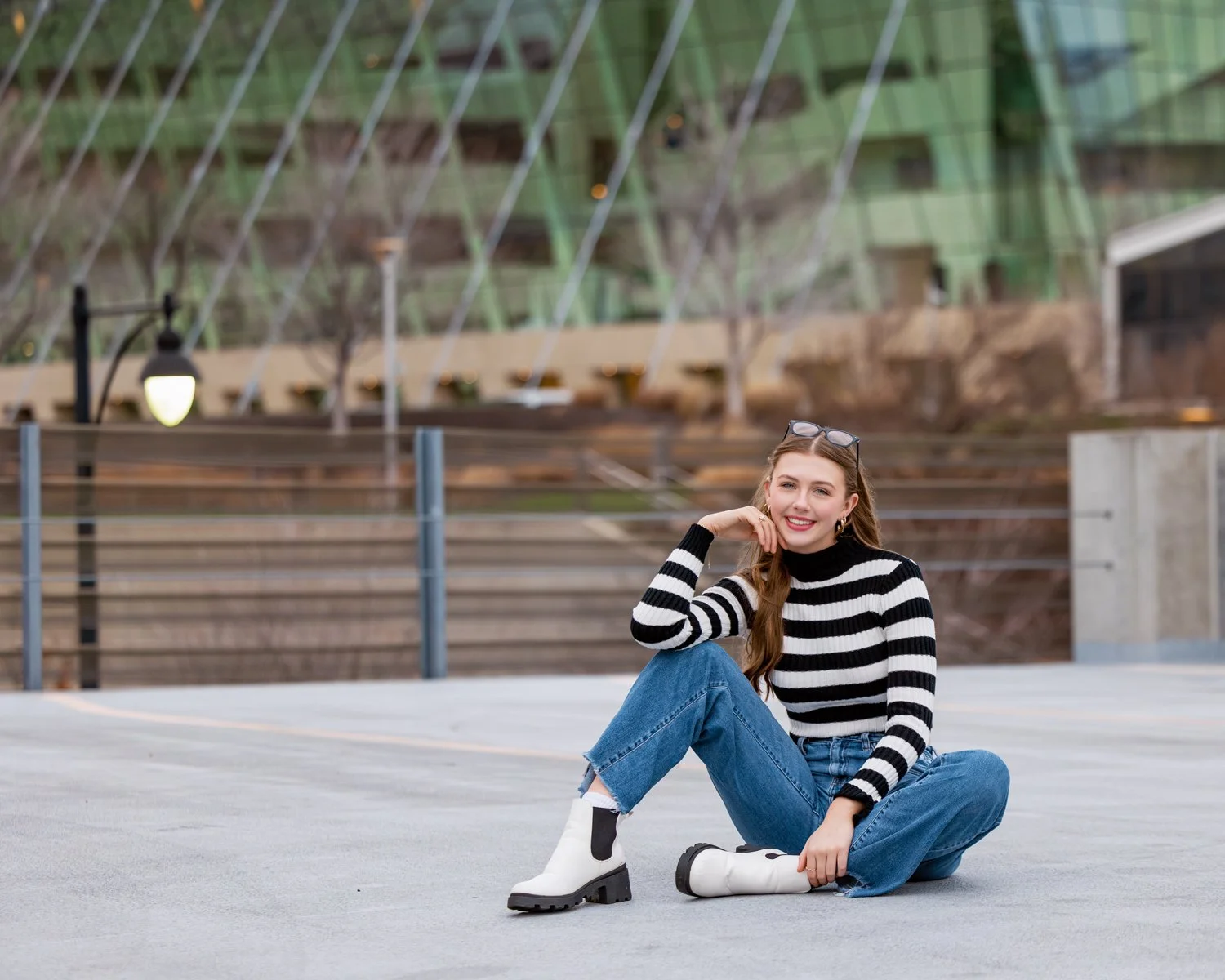 A high school senior girl, wearing a black and white striped turtleneck sweater, blue jeans, and white boots, sitting cross-legged with Kansas City Kauffman center in background.