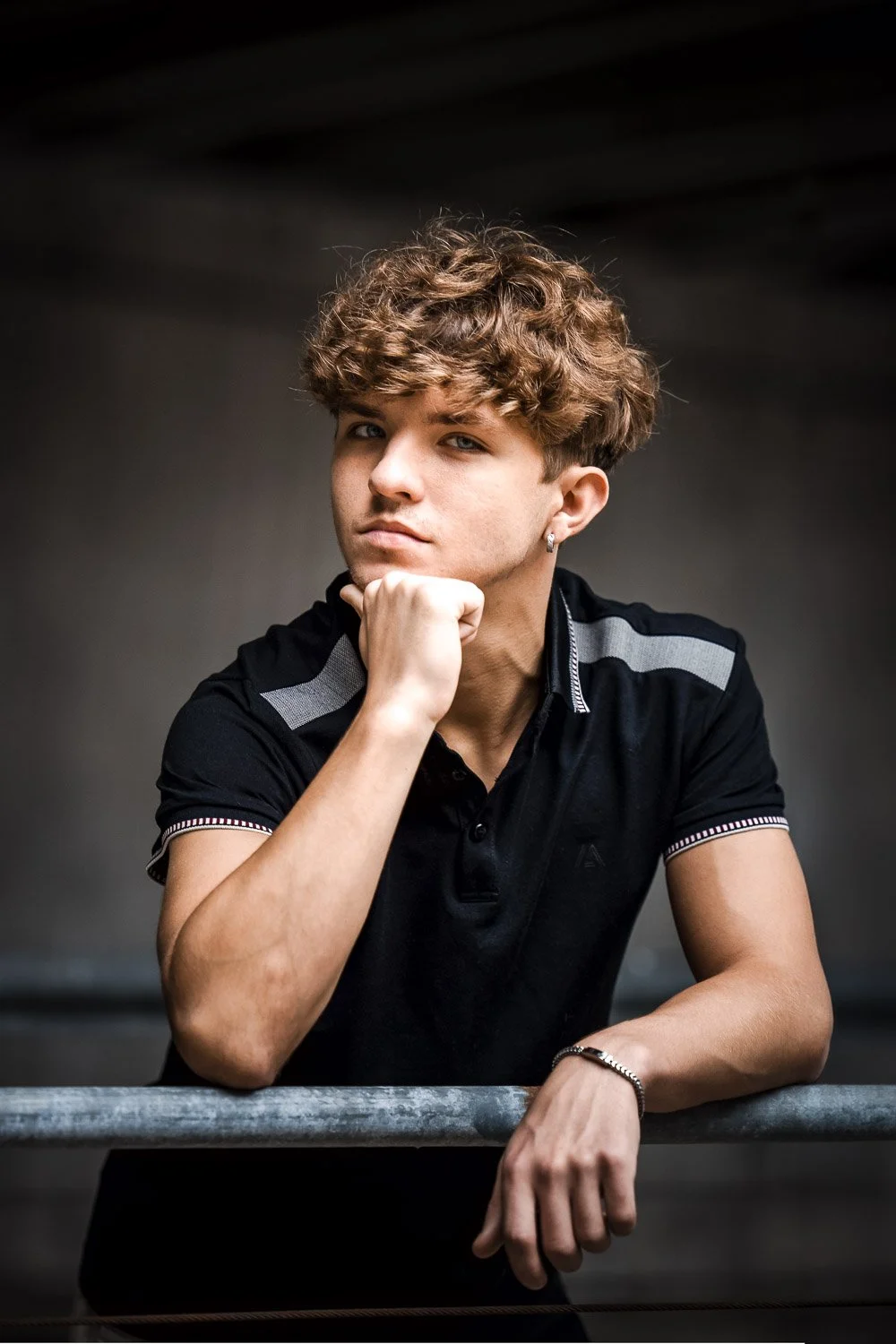 Editorial senior photo of a boy with curly hair, wearing a black polo shirt, resting his chin on his hand and looking at the camera, in a dimly lit setting outside Kansas City Union Station.