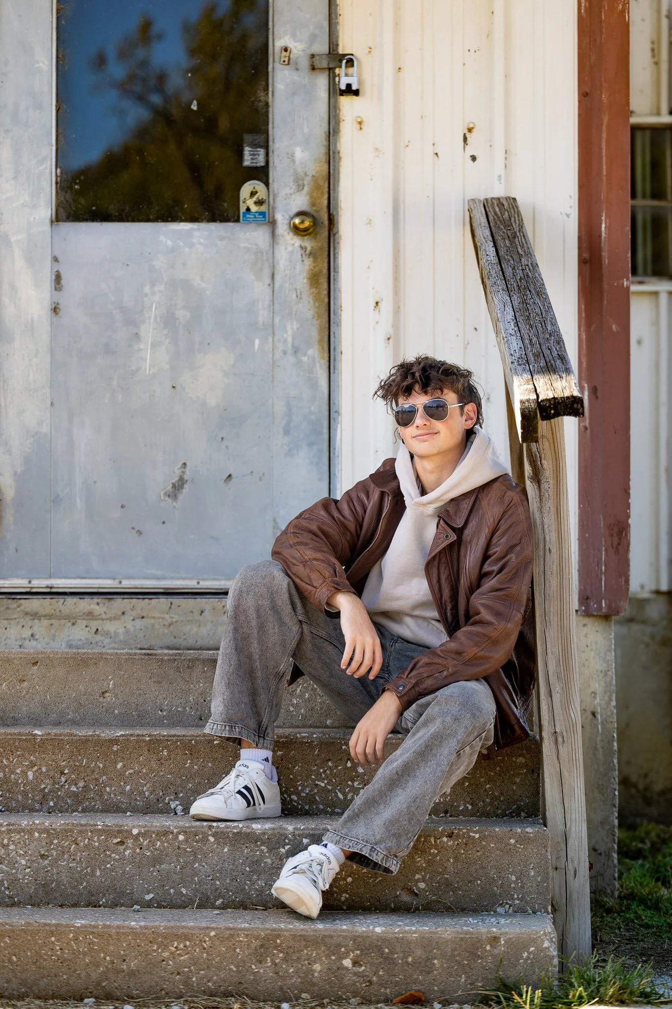 A high school senior boy sitting on outdoor concrete steps, wearing sunglasses, a leather jacket, a hoodie, gray jeans, and white sneakers, with a weathered wooden handrail next to him and a metal building in the background in Weston, MO.