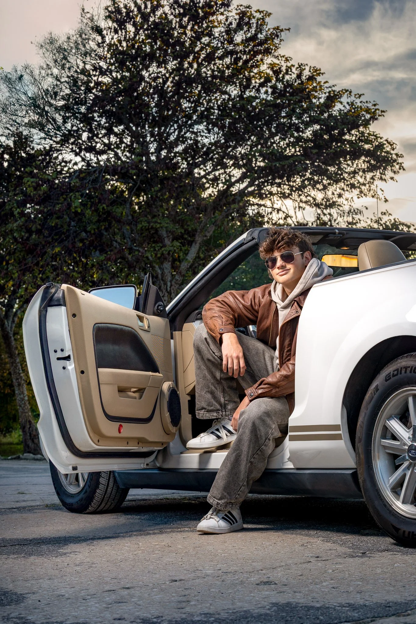 A high school senior sitting in the open door of a white convertible car, wearing sunglasses, a brown leather jacket, a hoodie, gray pants, and sneakers, with a tree and cloudy sky in the background.