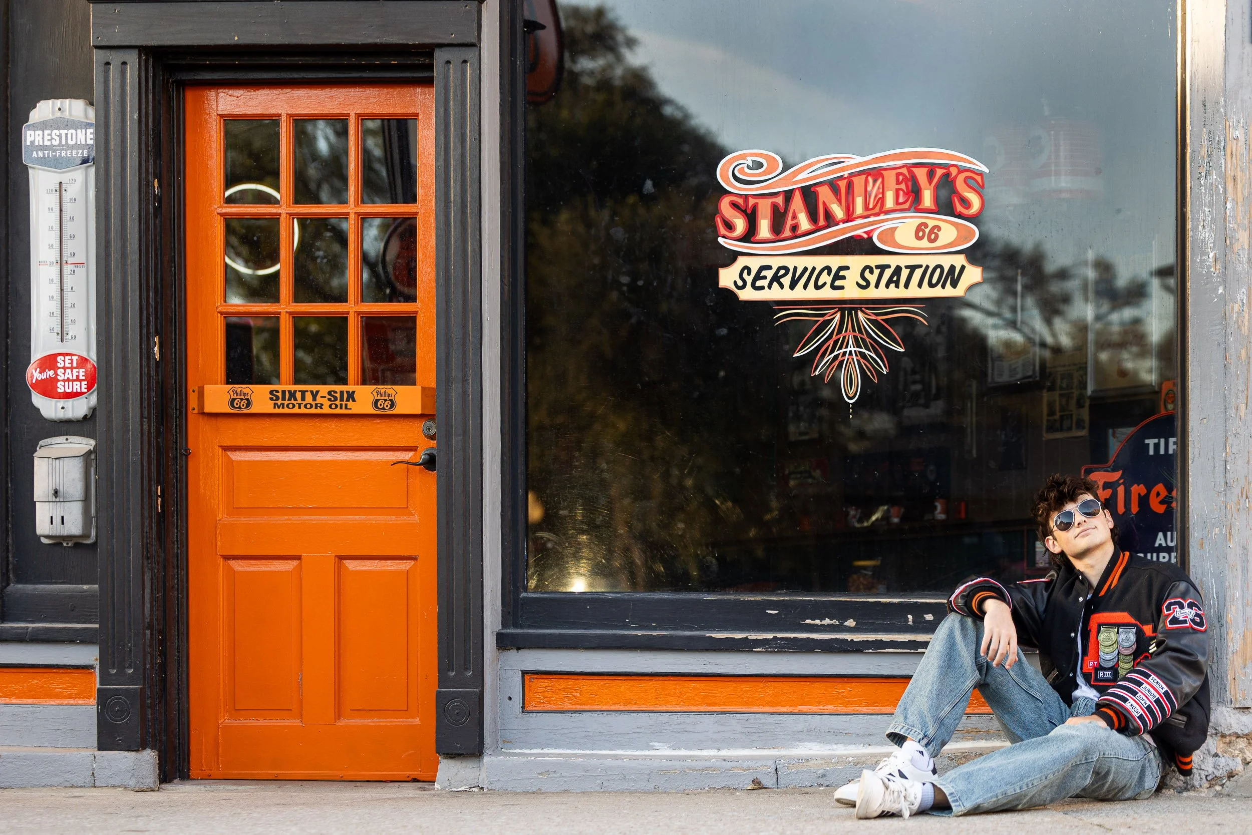 Senior boy at Weston Missouri gas station wearing sunglasses, a black varsity jacket with patches, and jeans sitting on the sidewalk