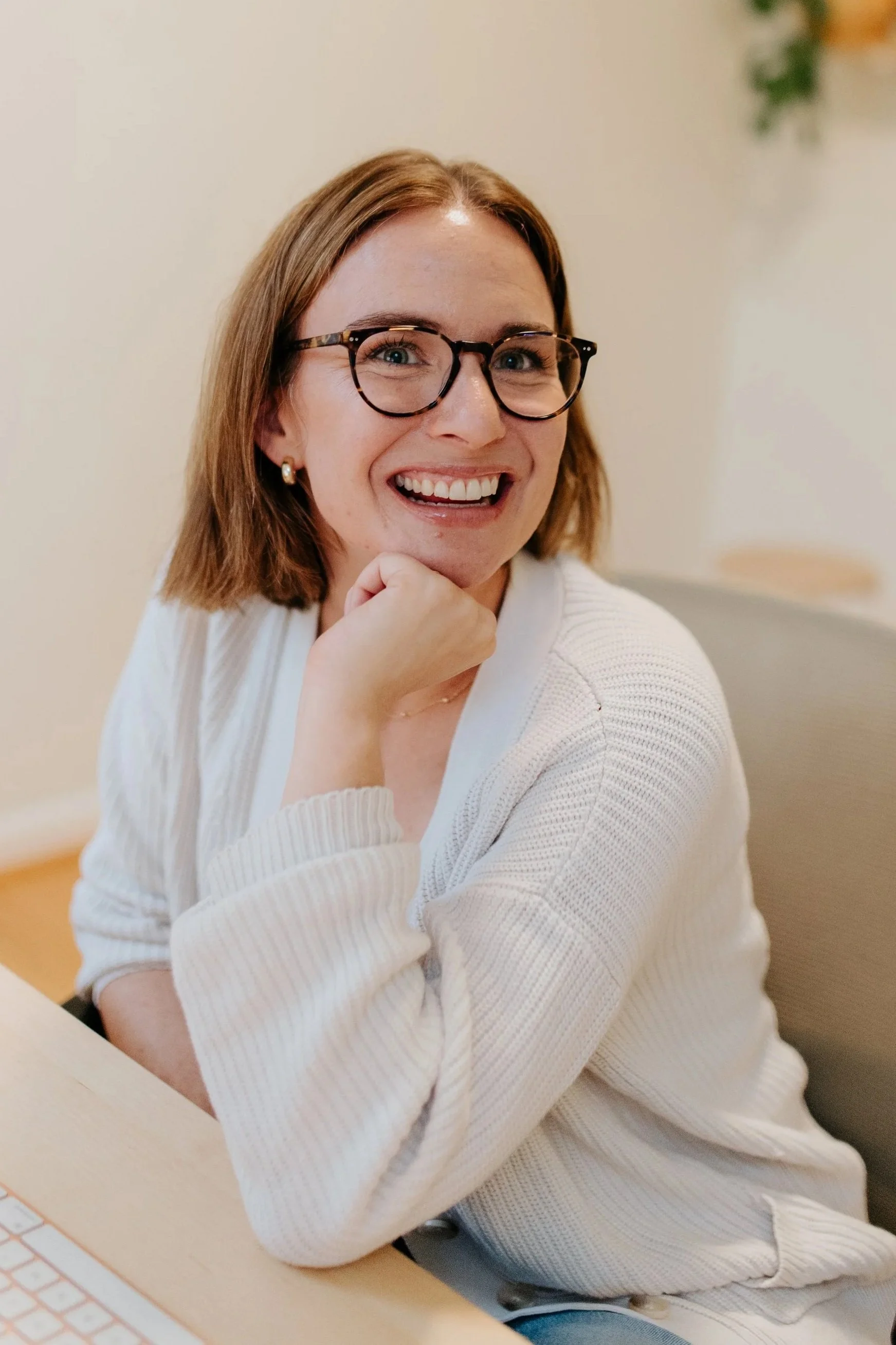 A woman with shoulder-length reddish hair, glasses, and earrings, smiling and leaning her chin on her hand, sitting at a desk with a keyboard in front of her, indoors with a light-colored wall background.