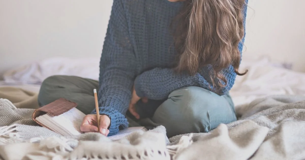 A picture of a woman in a blue sweater journaling in her bed. This image illustrates how to use this self compassion journaling template created by ellie miller lcsw-c, an online therapist in Maryland offering therapy to women with anxiety & couples