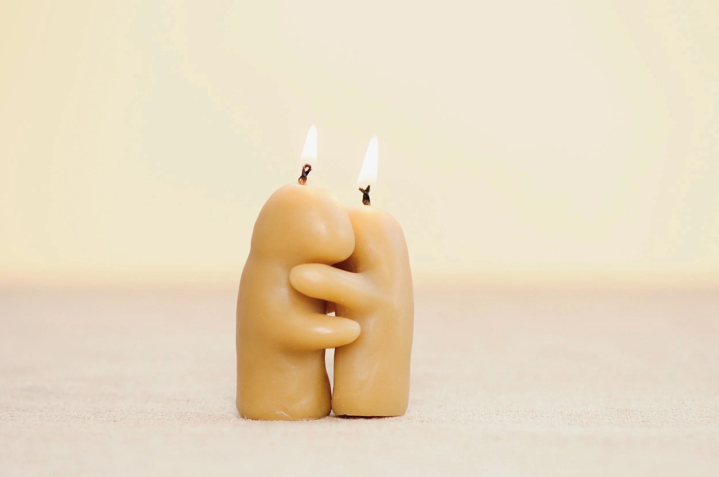 Two candles shaped like human figures holding each other, with one having a flame. The candles are beige and set against a plain background.