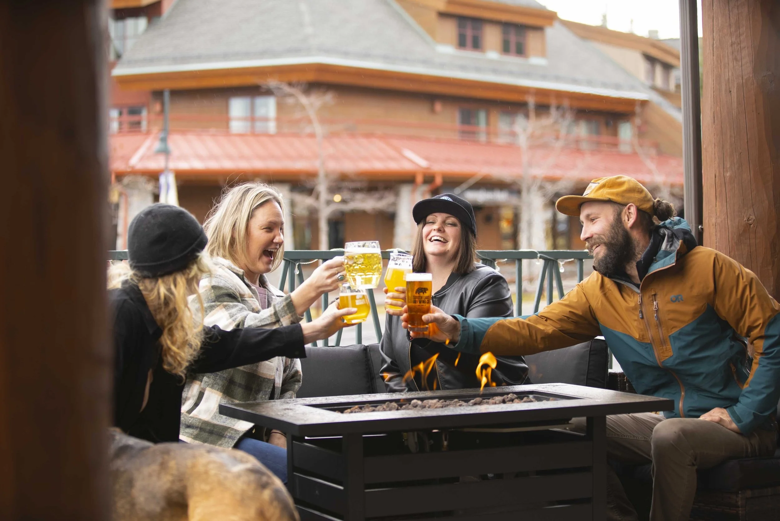 3 women and one man smile while doing a cheers with pints of beer around a firepit