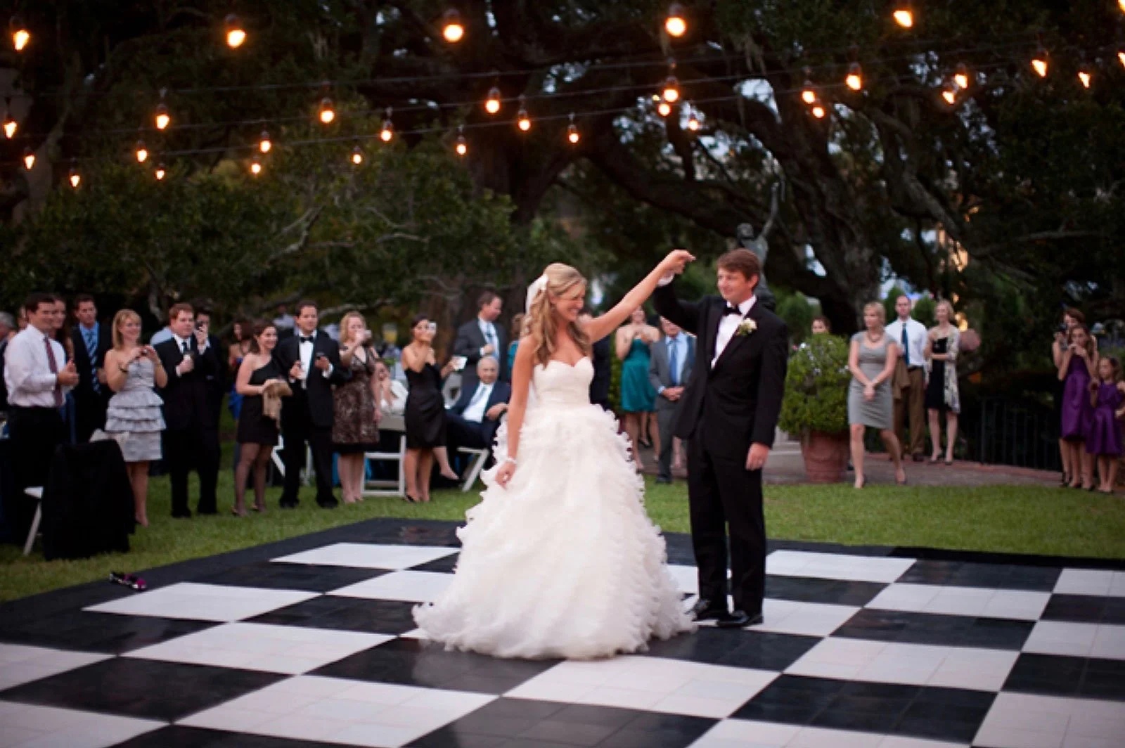 A bride and groom dancing on a checkered dance floor at an outdoor wedding reception, surrounded by guests, with string lights hanging above and large trees in the background.