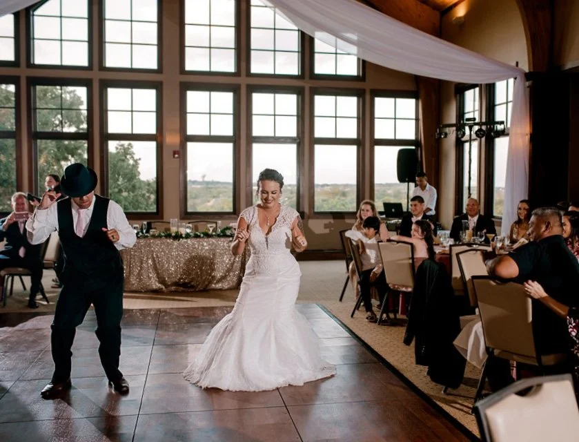 Bride dancing with a groom in a suit and hat at a wedding reception with seated guests watching.