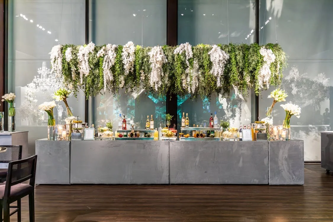 A decorated bar or buffet station with floral arrangements and bottles of alcohol, adorned with white and green flowers hanging above.
