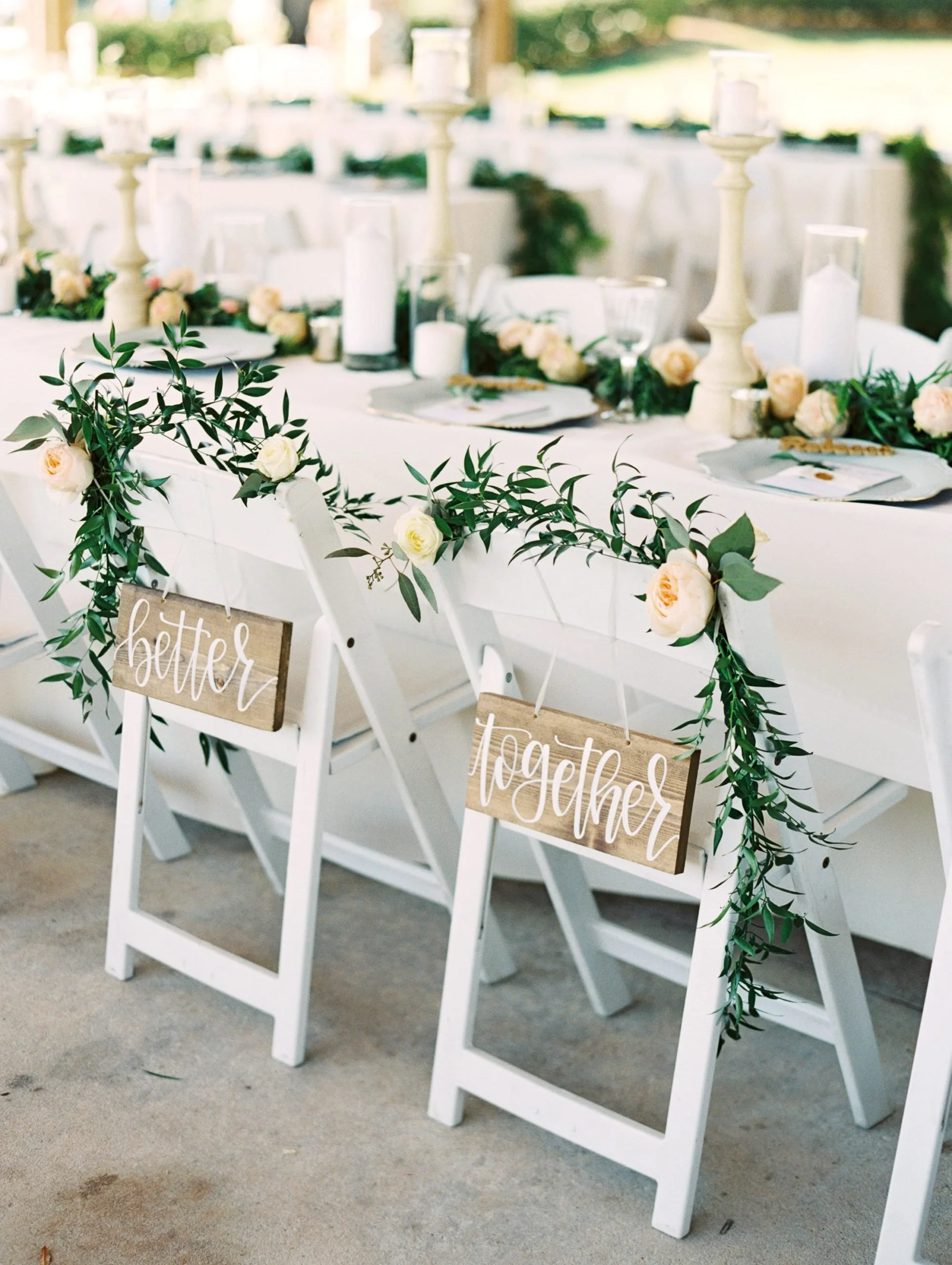 Wedding table decorated with white flowers, candles, and green foliage. Chairs have signs saying 'better together,' with greenery and roses attached.