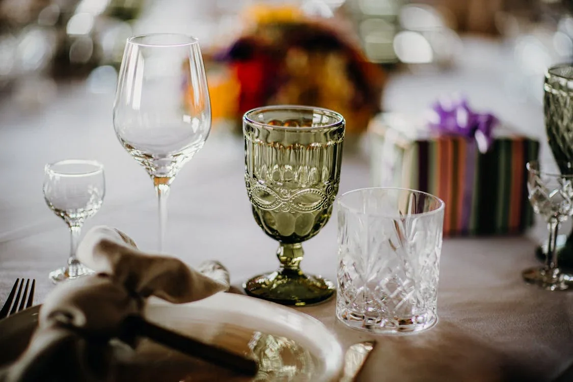 Elegant table setting with wine glasses, a gourd-shaped glass, a textured tumbler, a gold-colored goblet, and a wrapped gift box in the background
