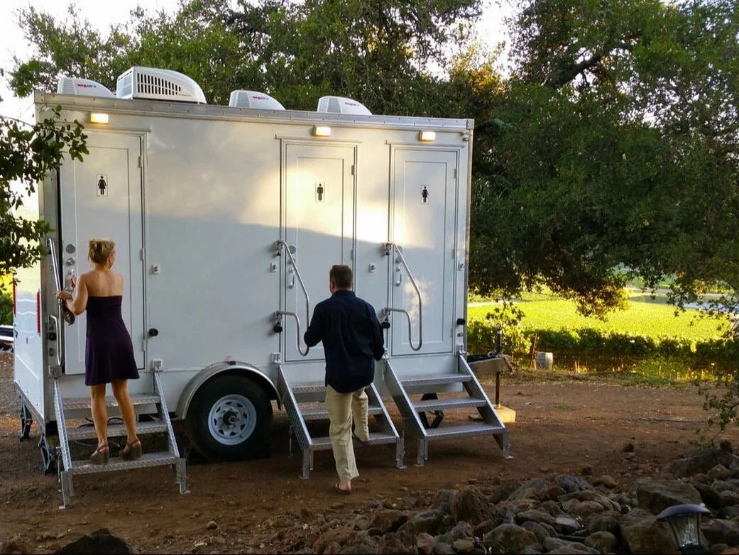 Public restroom trailer with three entry doors, each marked with a male or female sign, set outdoors on dirt ground with trees in the background, and two people approaching it.