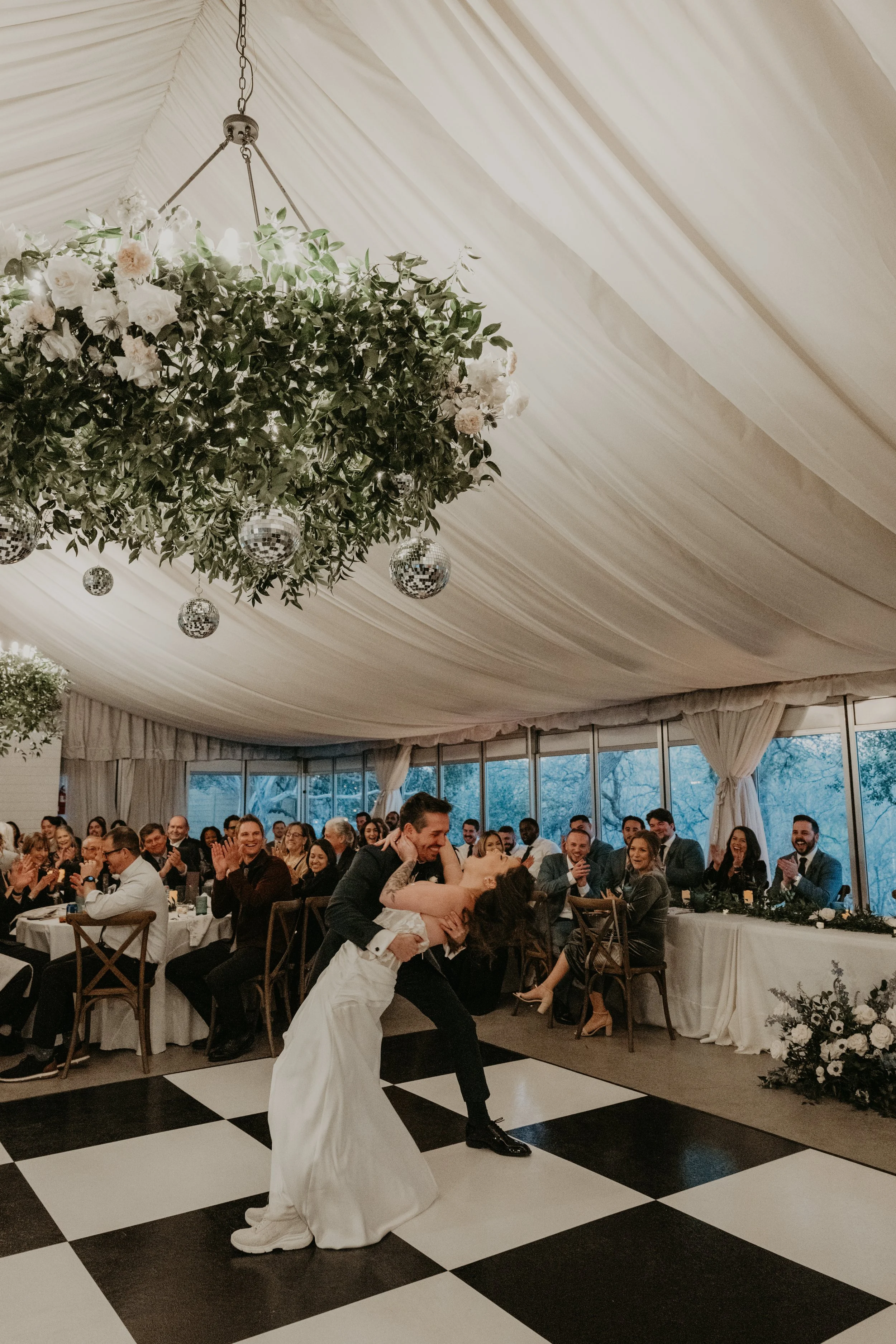 A bride and groom dancing on a black-and-white checkered dance floor at their wedding reception, surrounded by seated guests in a decorated indoor venue.