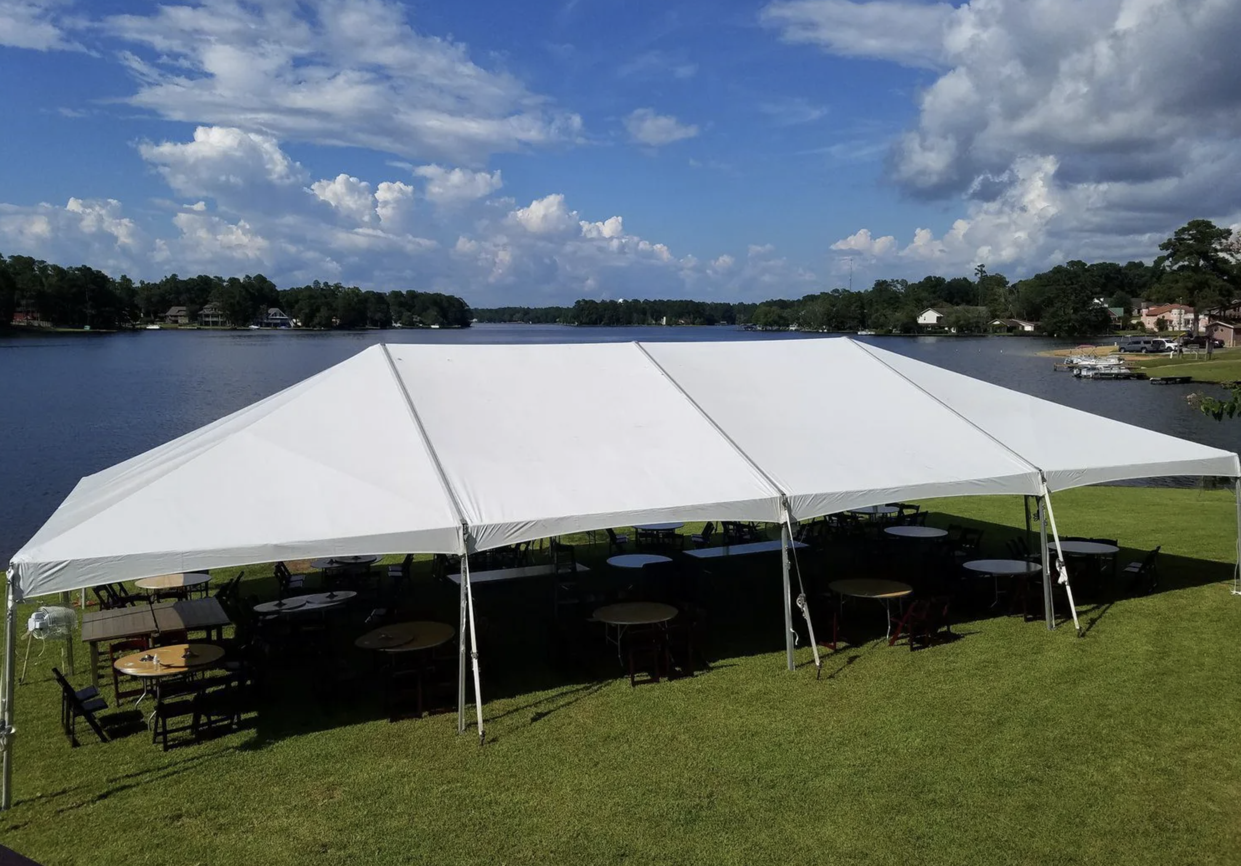 White event tent with tables and chairs set up on a grassy area near a lake under a partly cloudy sky.