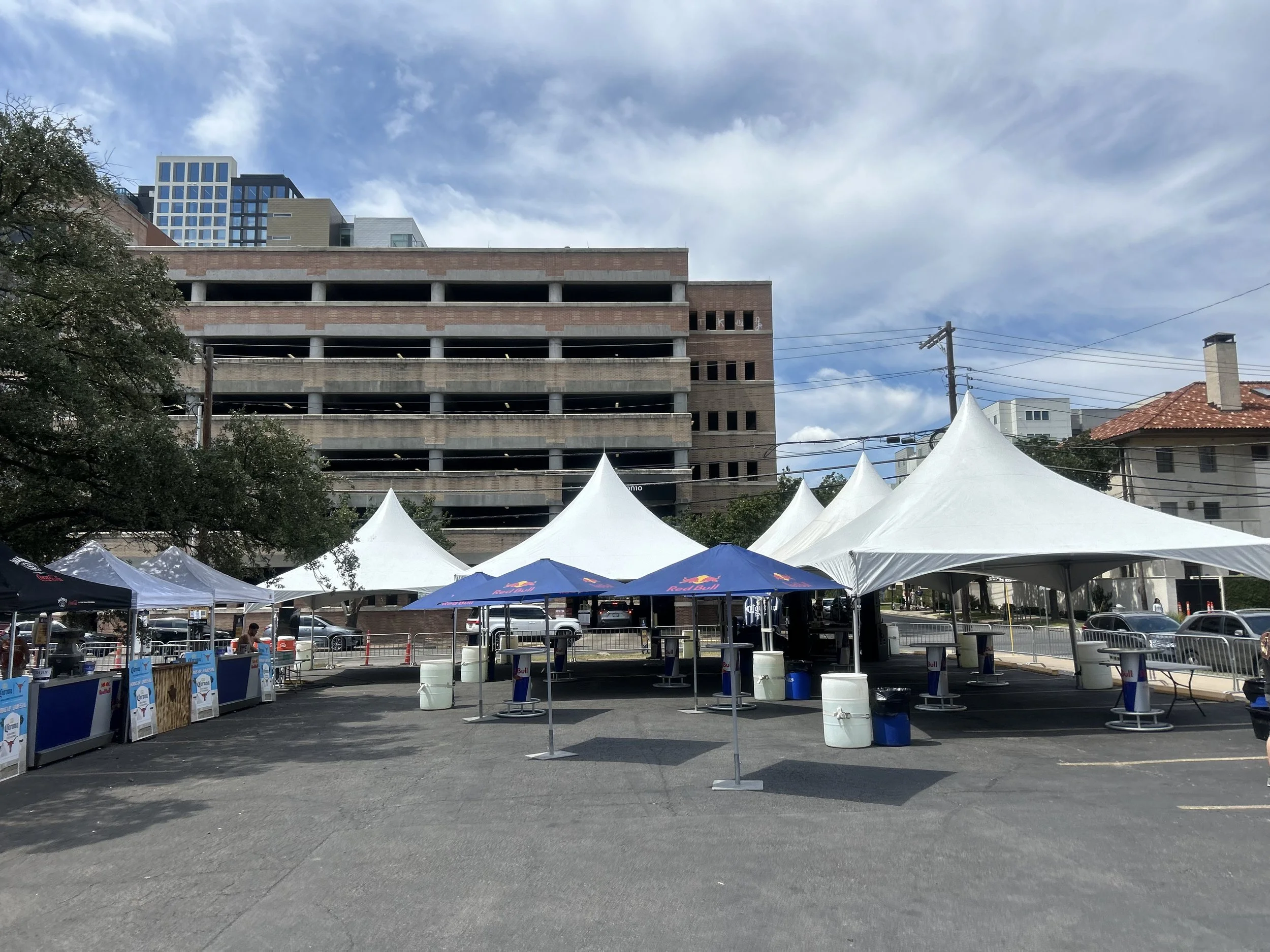 Outdoor event setup with large white tents and smaller tents with umbrellas, tables, and barricades in a parking lot, with an unfinished multi-story building and residential houses in the background under a partly cloudy sky.