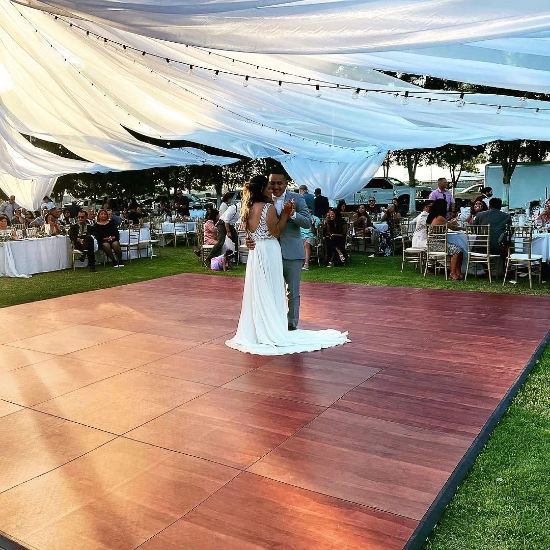 A bride and groom dancing on a wooden dance floor under a canopy at their outdoor wedding reception, with guests seated at tables in the background.