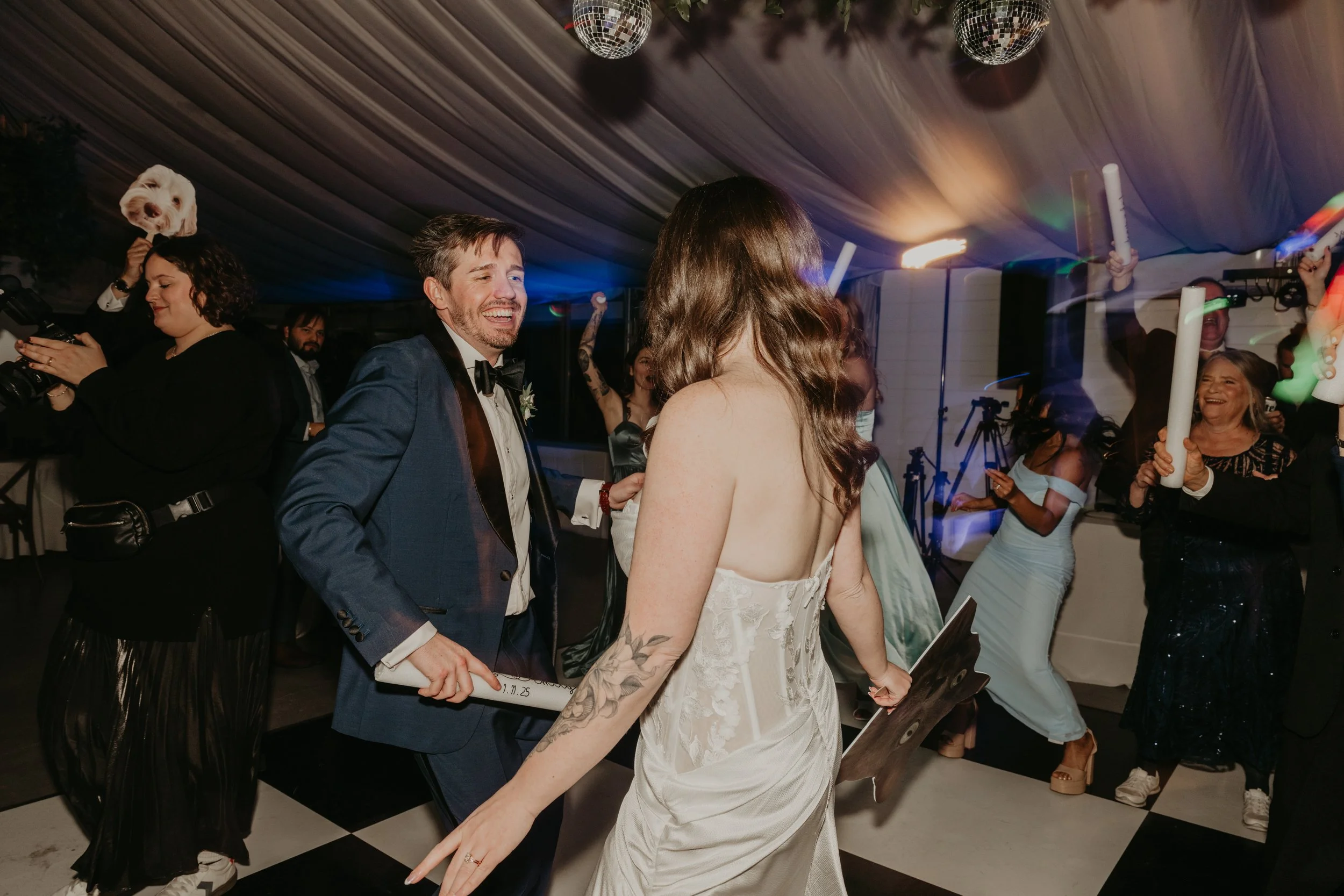 A wedding reception with people dancing, holding glow sticks, and celebrating under a decorated tent ceiling with disco balls and lighting.