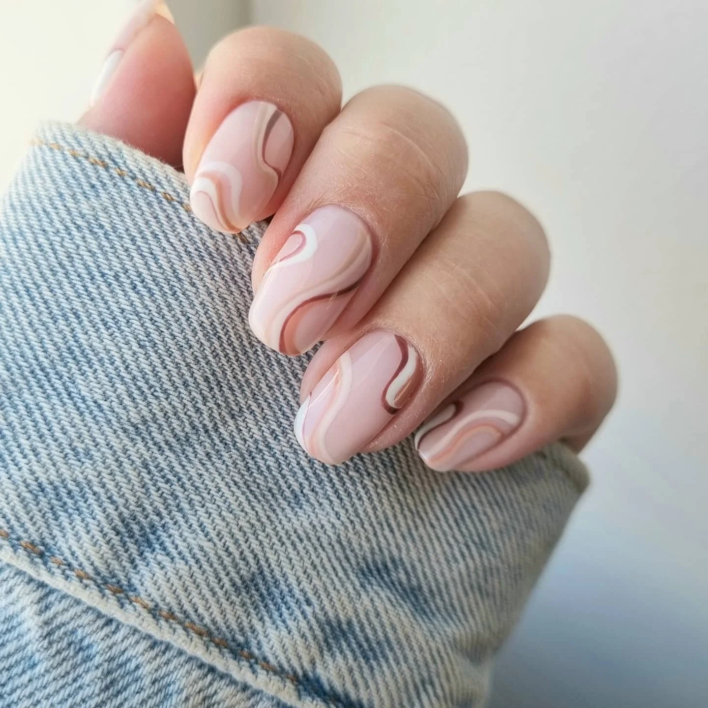 Nærbillede af manicure med lyserøde negle med hvide og brune bølgeformede mønstre, en person bruger lyseblå jeans.