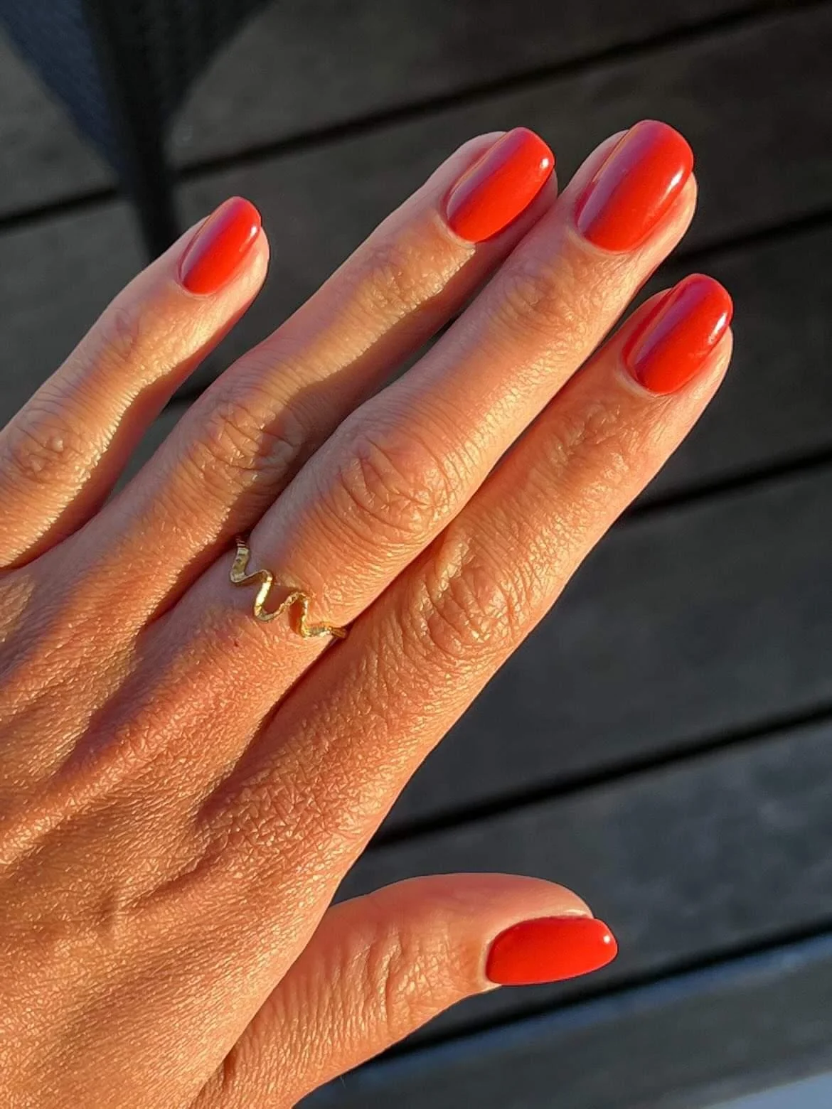 Hand med rødne negle og et gold ring med zigzag design, mod en mørk træbaggrund.
