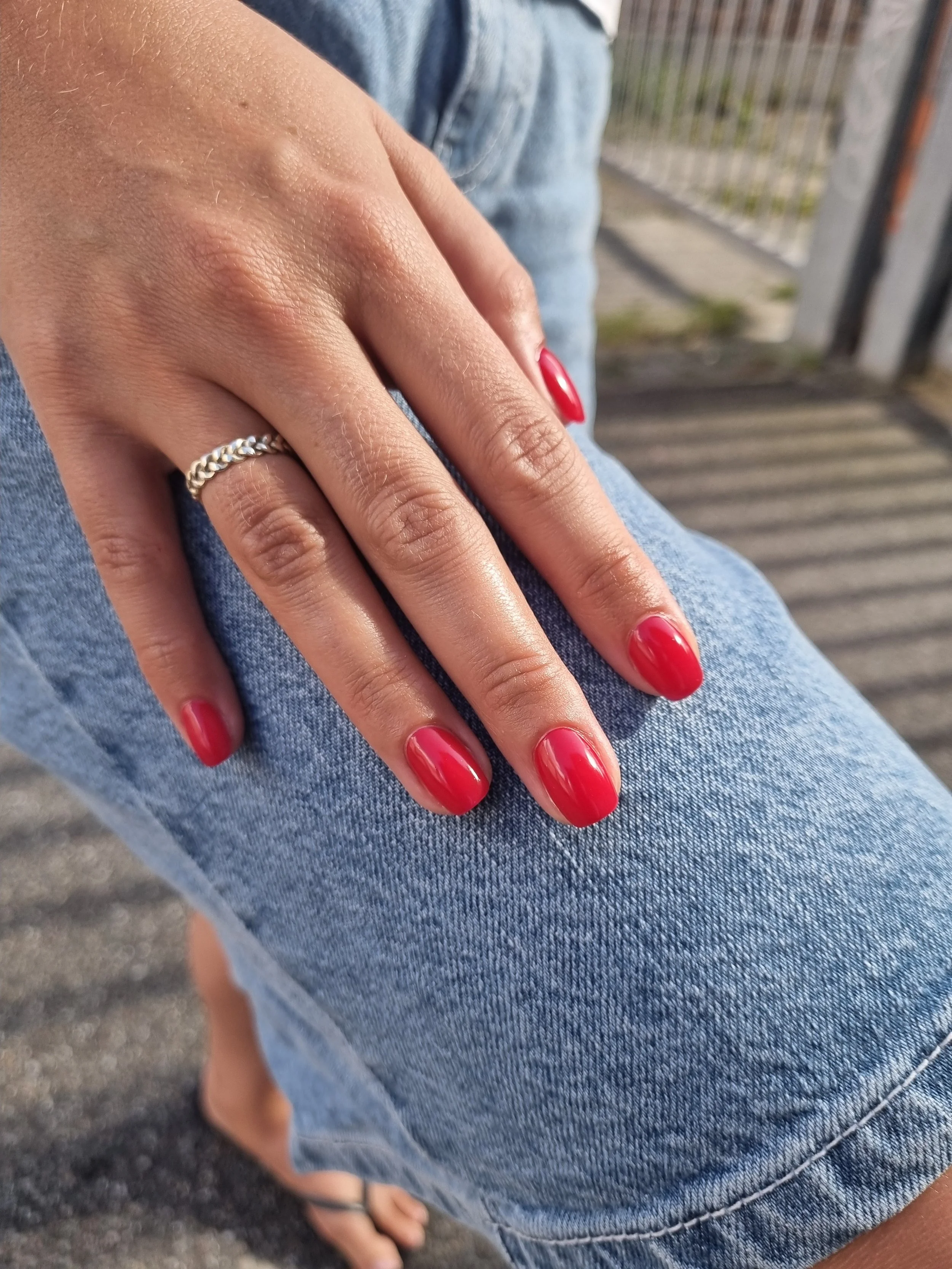 Nærbillede af en hånd med rødt neglelak, et sølvring på pegefingeren, person er iført blå denim jeans, og der er udendørs, med en flisetjæppe og et udendørs gelænder i baggrunden.