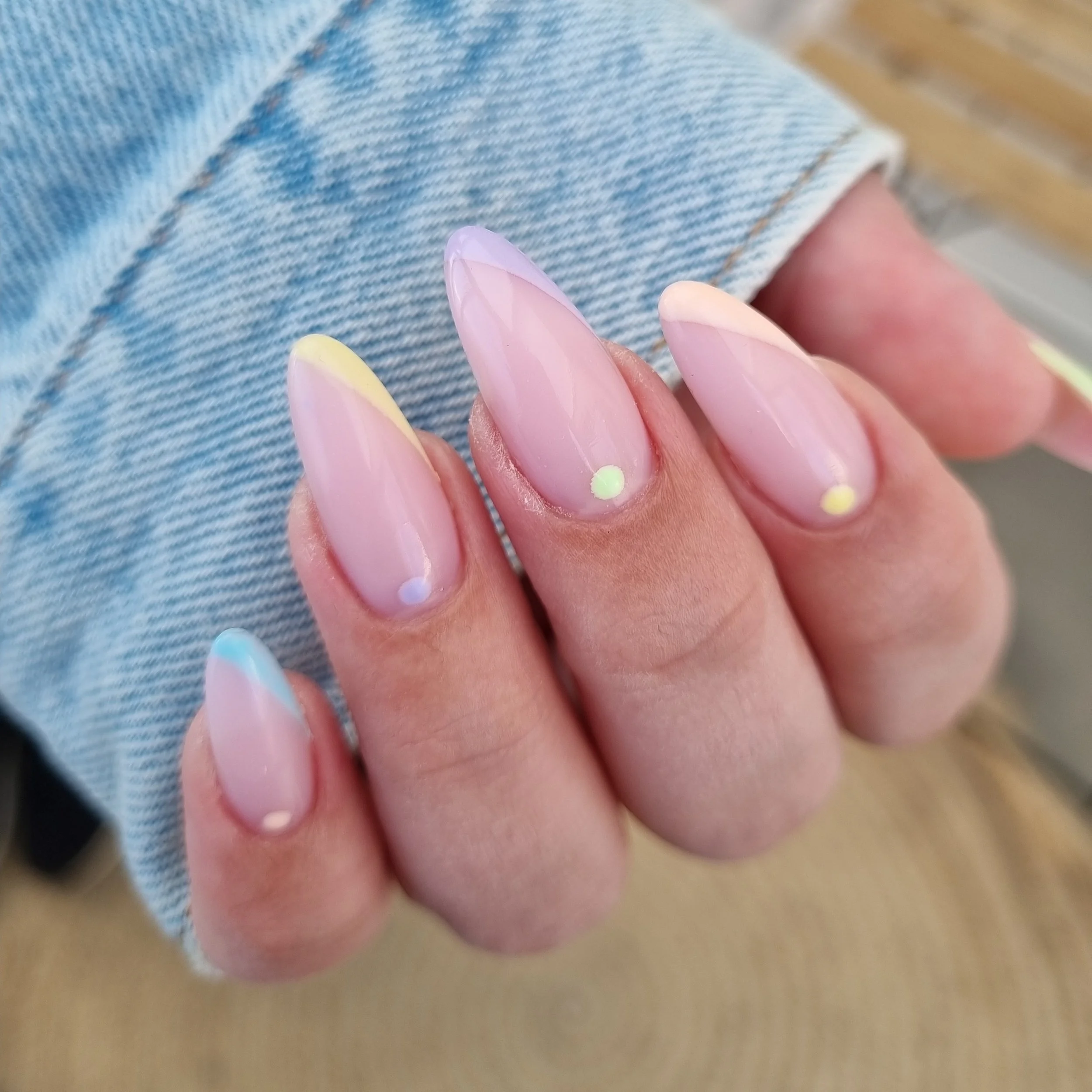 Nærbillede af en persons hånd med lang, oval formede negle, der har en lakering i pastelfarver med små farvede prikker.