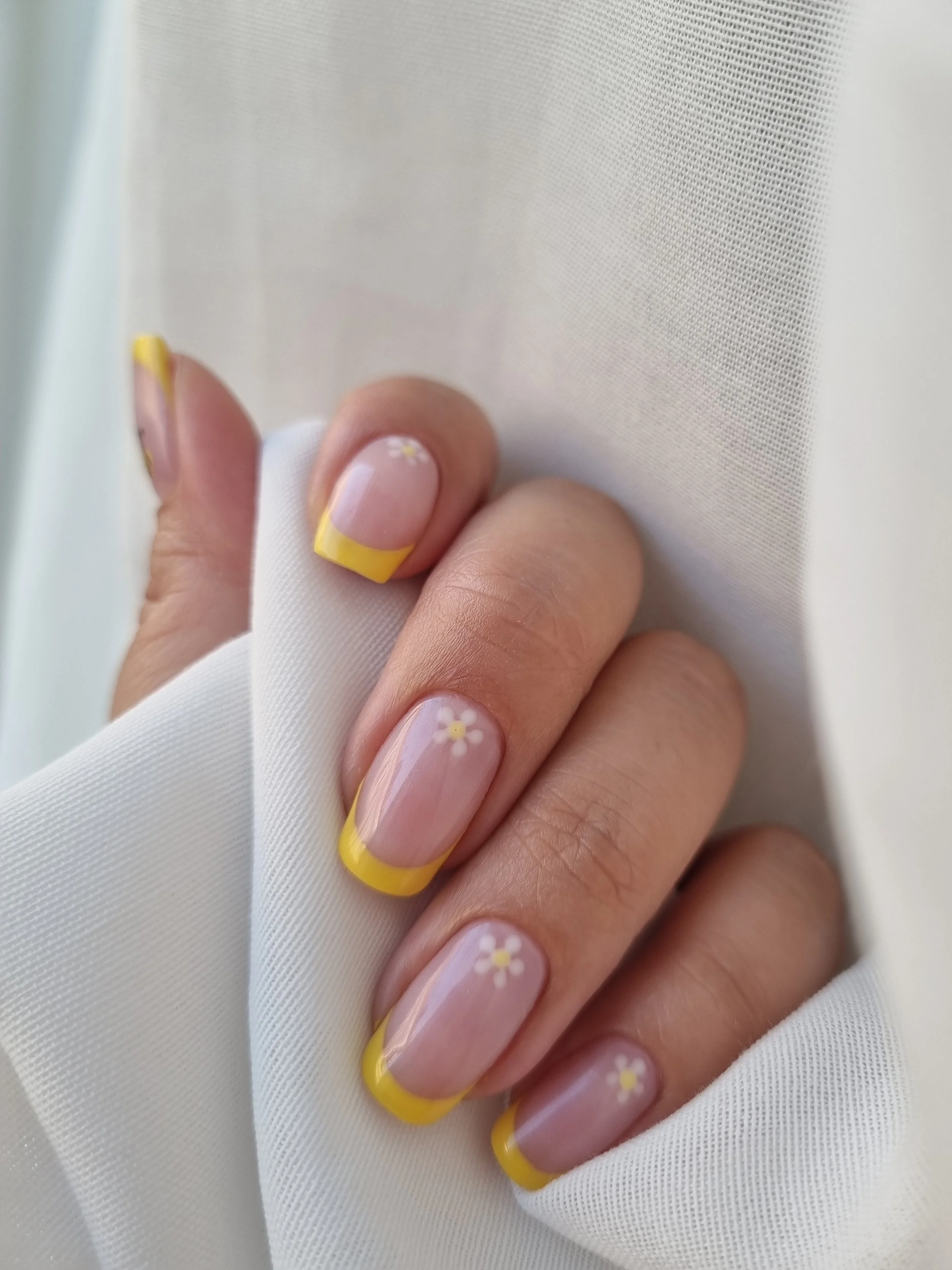 Nærbillede af en hånd med neglelækker i lyserød med gule spidser og små hvide blomster. Hånden holder et stykke hvidt stof.