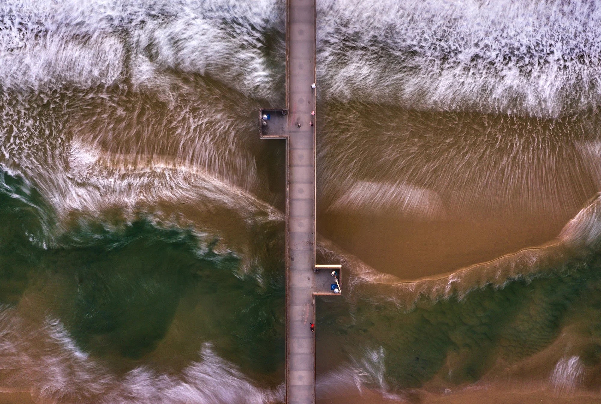 Aerial view of a pier extending into the ocean, with waves crashing on both sides.