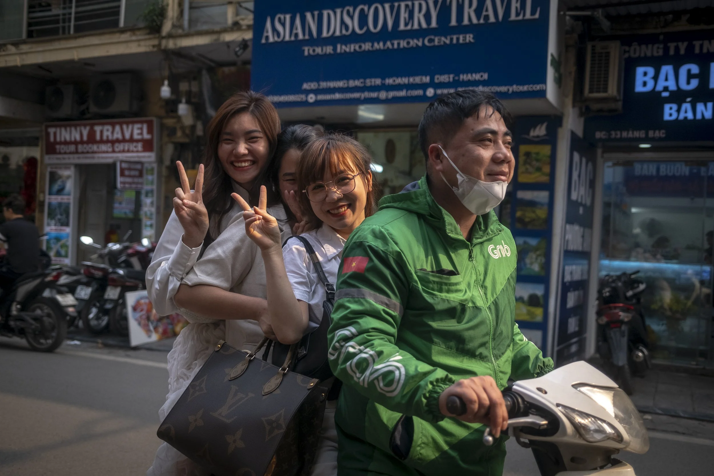 Four women riding on a motorcycle driven by a man wearing a green jacket and face mask, smiling and making peace signs in an urban street with travel agency signs in the background.