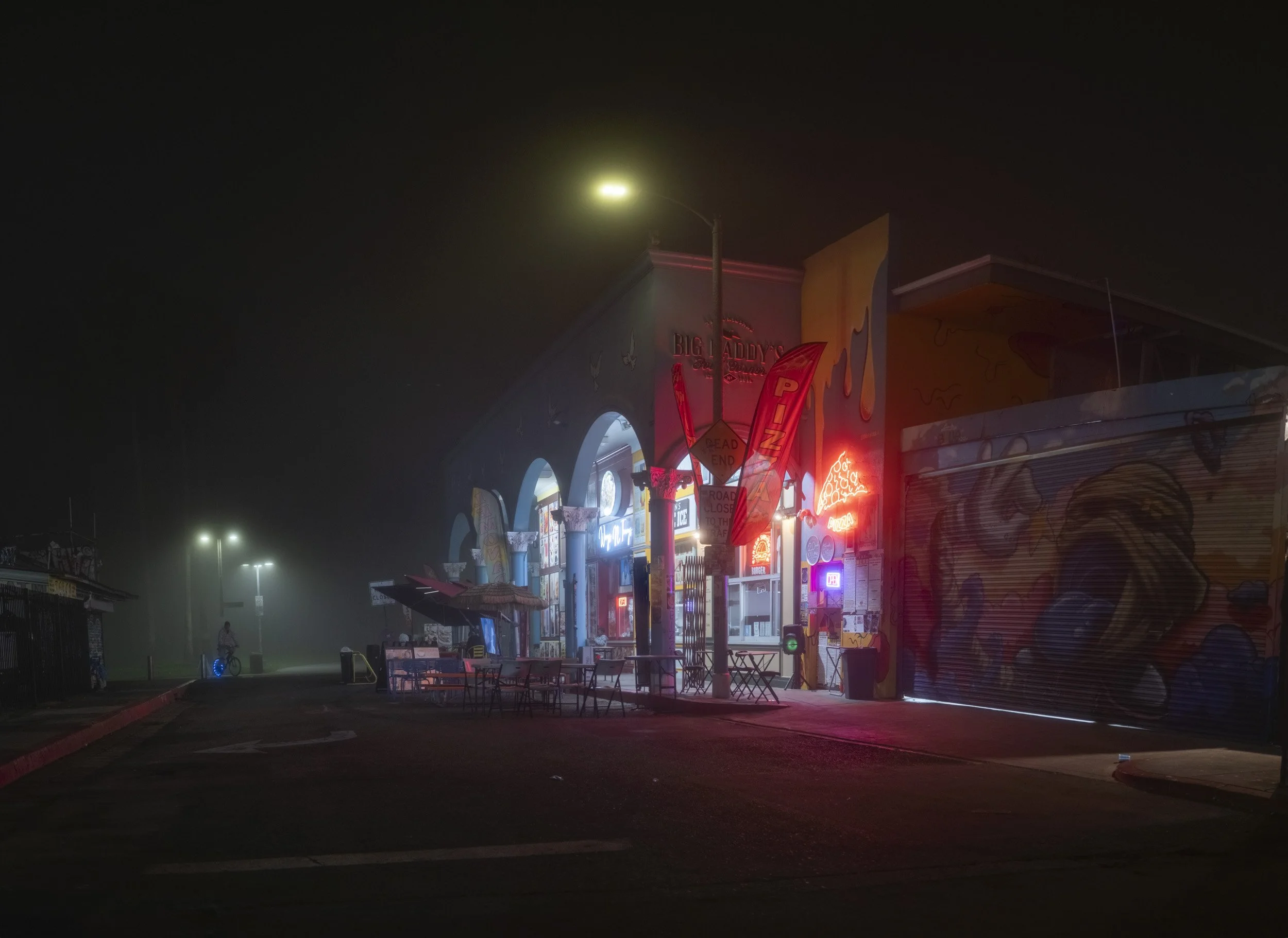 Nighttime scene of a closed colorful storefront with neon signs and painted walls, illuminated by streetlights and fog.