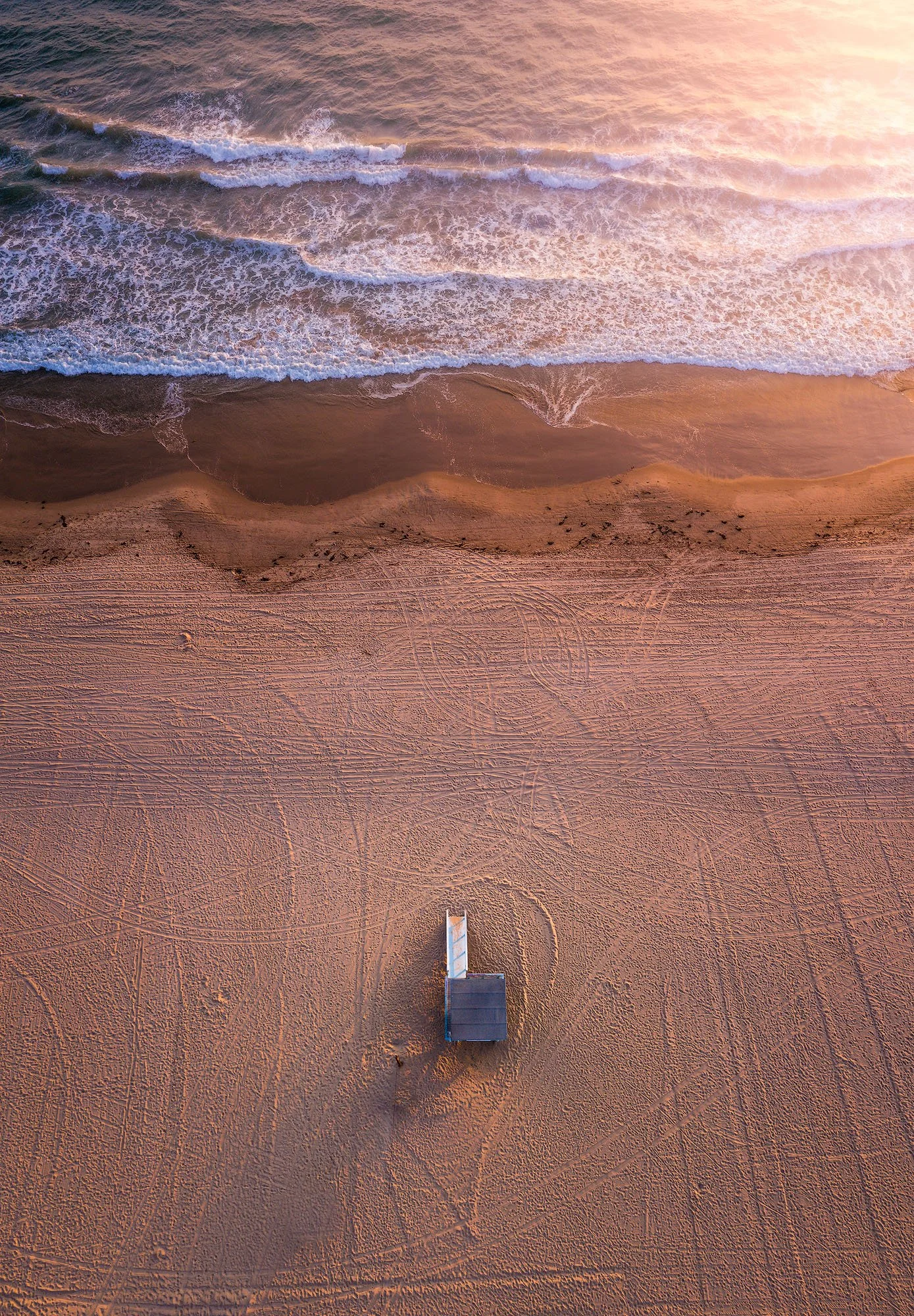 An aerial view of a sandy beach at sunrise with gentle ocean waves, and a small beach hut in the center, casting a shadow on the sand.