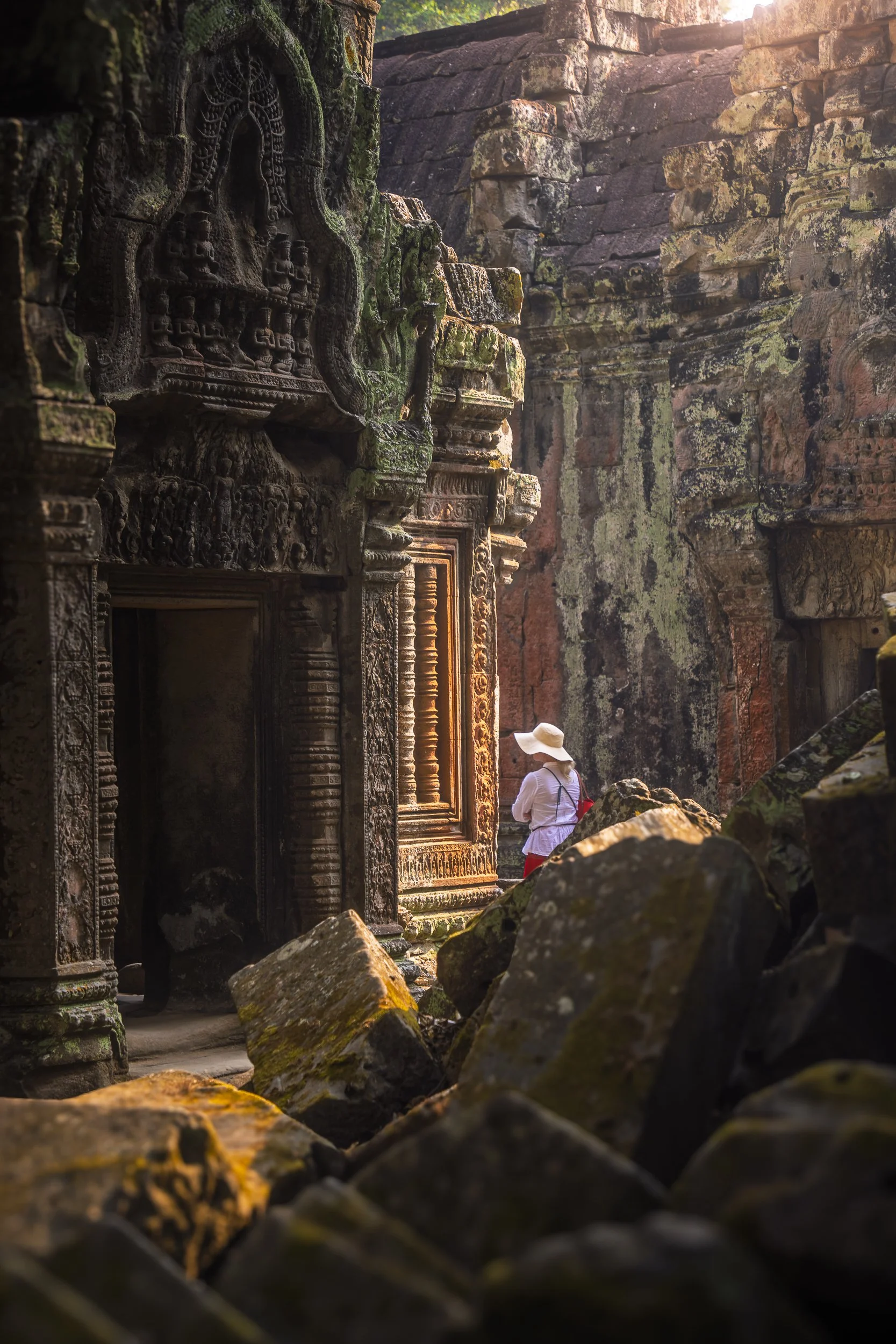A person wearing a white dress and wide-brimmed sun hat standing among ancient stone ruins with intricate carvings, surrounded by moss-covered stones and illuminated by warm sunlight.