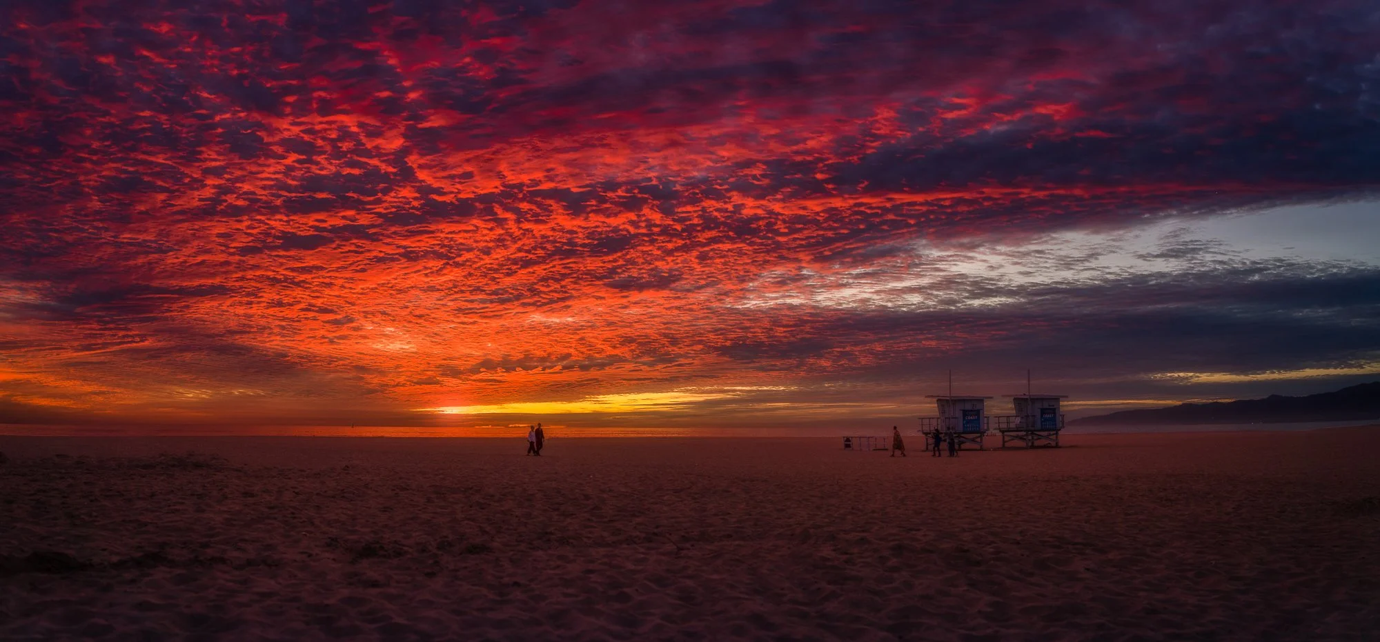 A vibrant sunset over the beach with a colorful sky filled with shades of red, orange, and purple, and a few people walking near lifeguard towers.