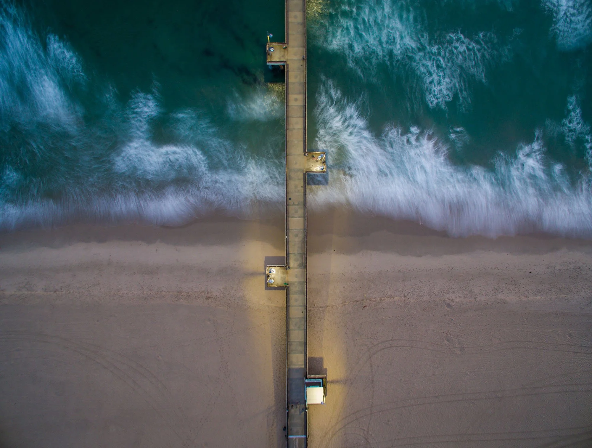 Aerial view of a beach with ocean waves, sandy shoreline, and a long pier extending into the water, illuminated by lights.