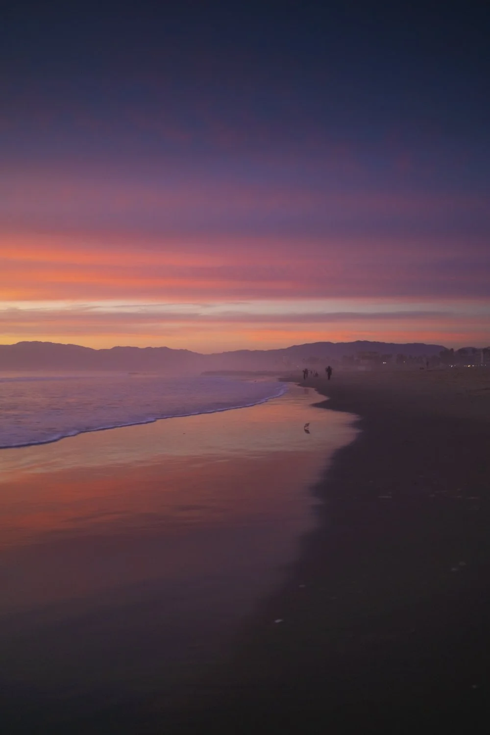 Sunset over the beach with colorful sky, silhouettes of people walking along the shoreline, and calm water reflecting the sky's hues.