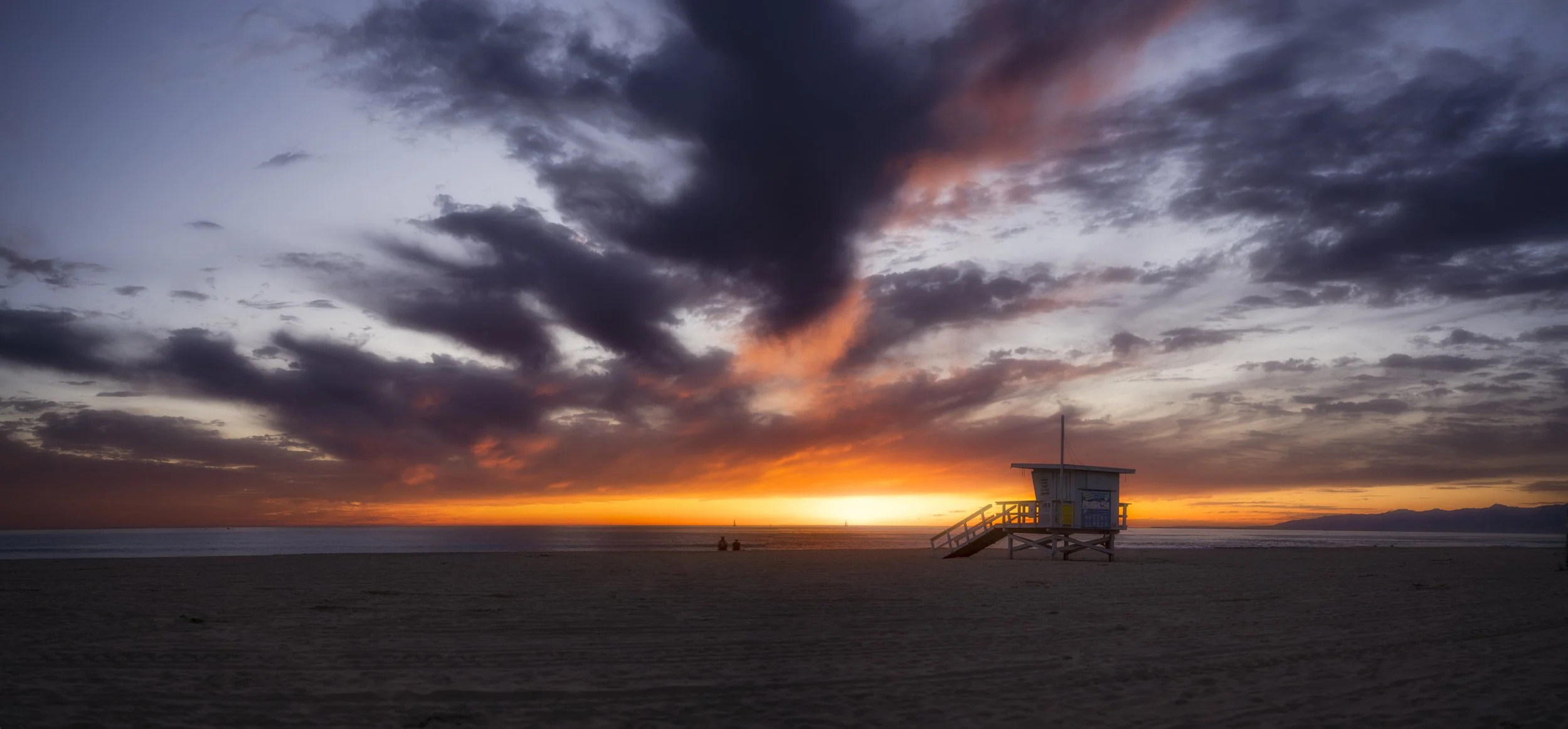 Beach scene at sunset with colorful clouds, a lifeguard tower on the right, and a few people sitting on the sand, with sailboats in the distance.