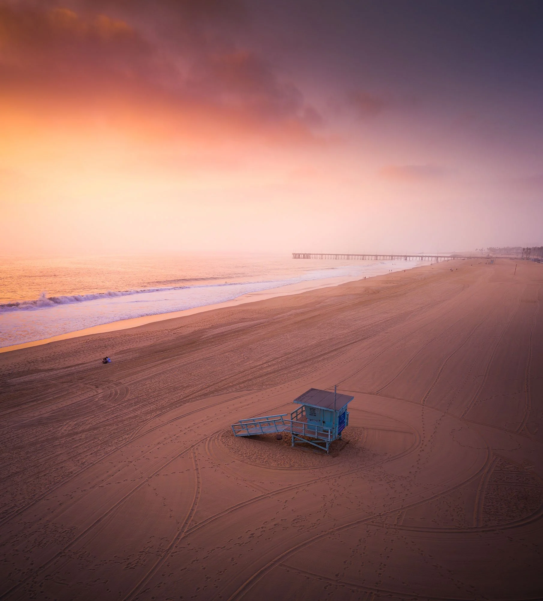 Empty sandy beach during sunset with a lifeguard tower in the foreground and a pier stretching into the ocean in the background.