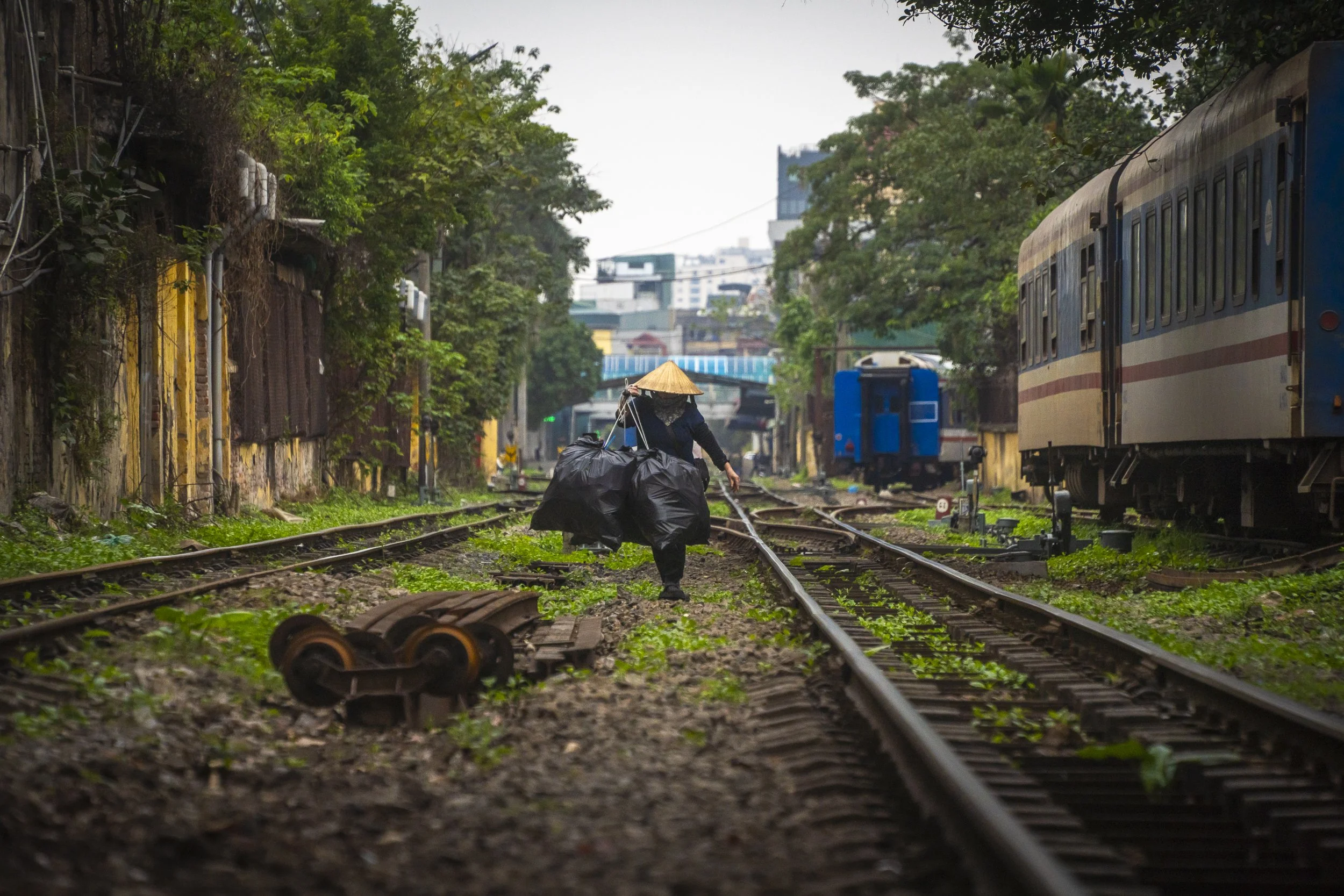 A person wearing a conical hat and black clothing walking along railway tracks, carrying large black bags and holding a walking stick, with green foliage on either side and train cars in the background.